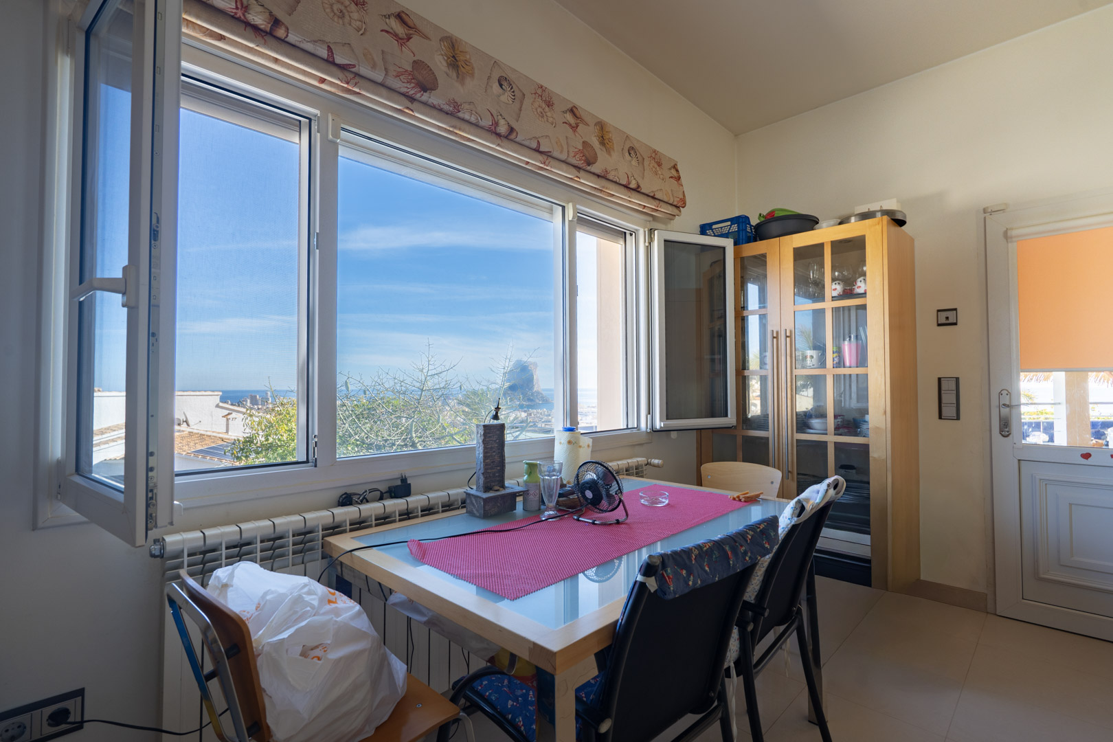Dining area featuring a large window with direct views of the Peñón de Ifach. Includes a glass-top wooden table, a display cabinet, and radiator heating.
