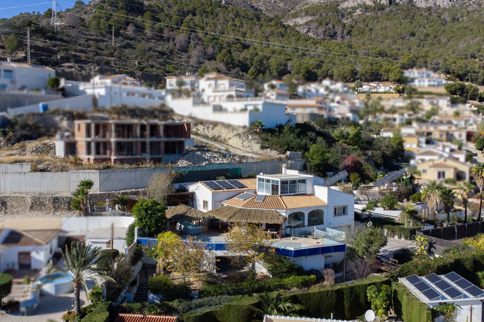Elevated view of the multi-level villa featuring rooftop solar panels, a glass-enclosed upper terrace, and expansive outdoor areas with thatched pergolas on a hillside.