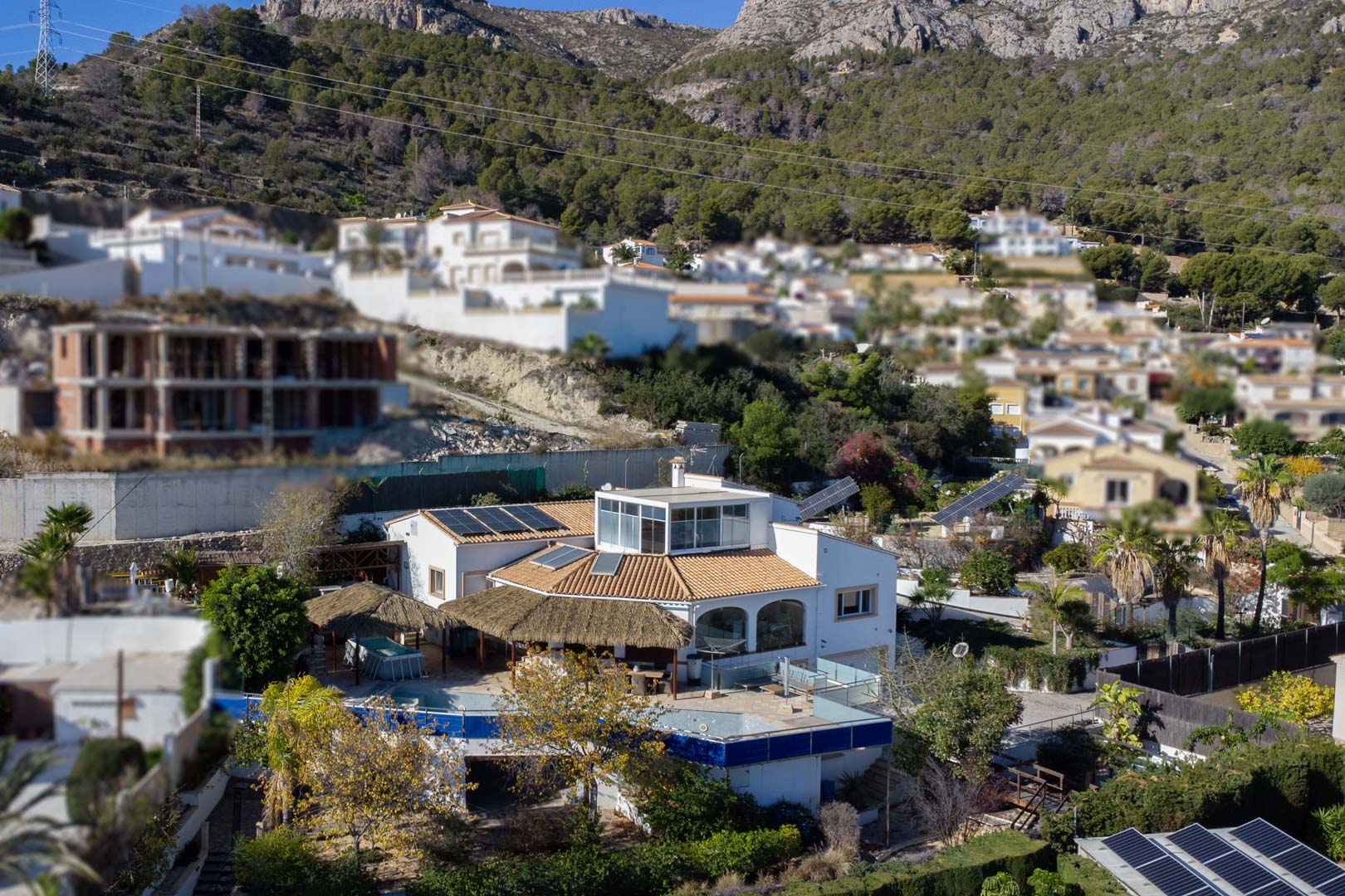 Elevated view of the villa featuring solar panel systems, a glass-enclosed upper terrace, and a pool area with thatched pergolas near the mountains.