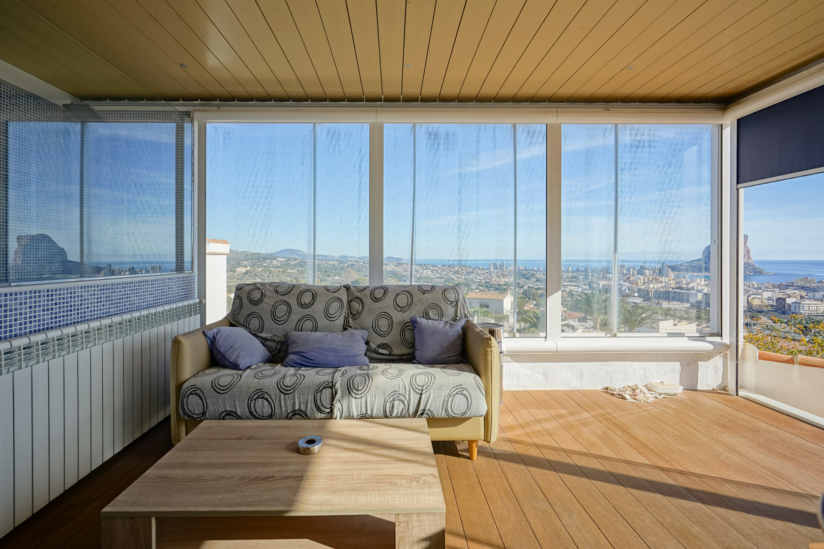 Enclosed terrace featuring glass curtains, wood-plank flooring, and panoramic views of the Calpe skyline and Peñón de Ifach. Includes a paneled ceiling and lounge area.