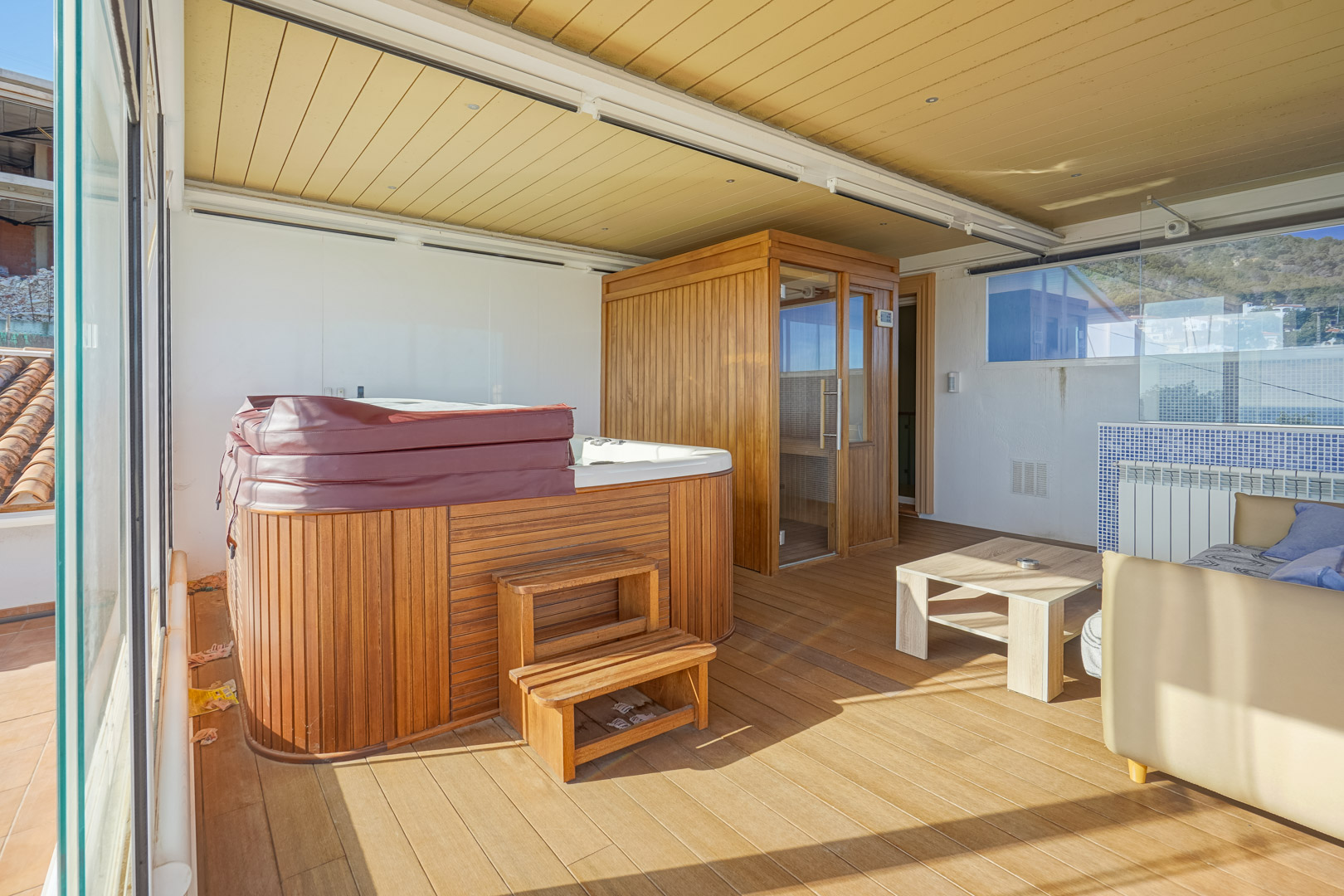 Enclosed upper terrace featuring a wood-paneled jacuzzi, a private sauna, and timber decking, all protected by glass curtains and a structured ceiling with recessed lighting.