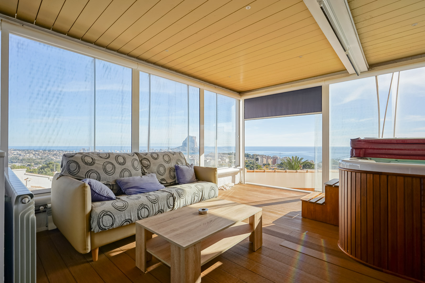 Enclosed upper terrace featuring a wood-paneled jacuzzi, wooden flooring, and floor-to-ceiling glass walls with views of the Peñón de Ifach and the sea.