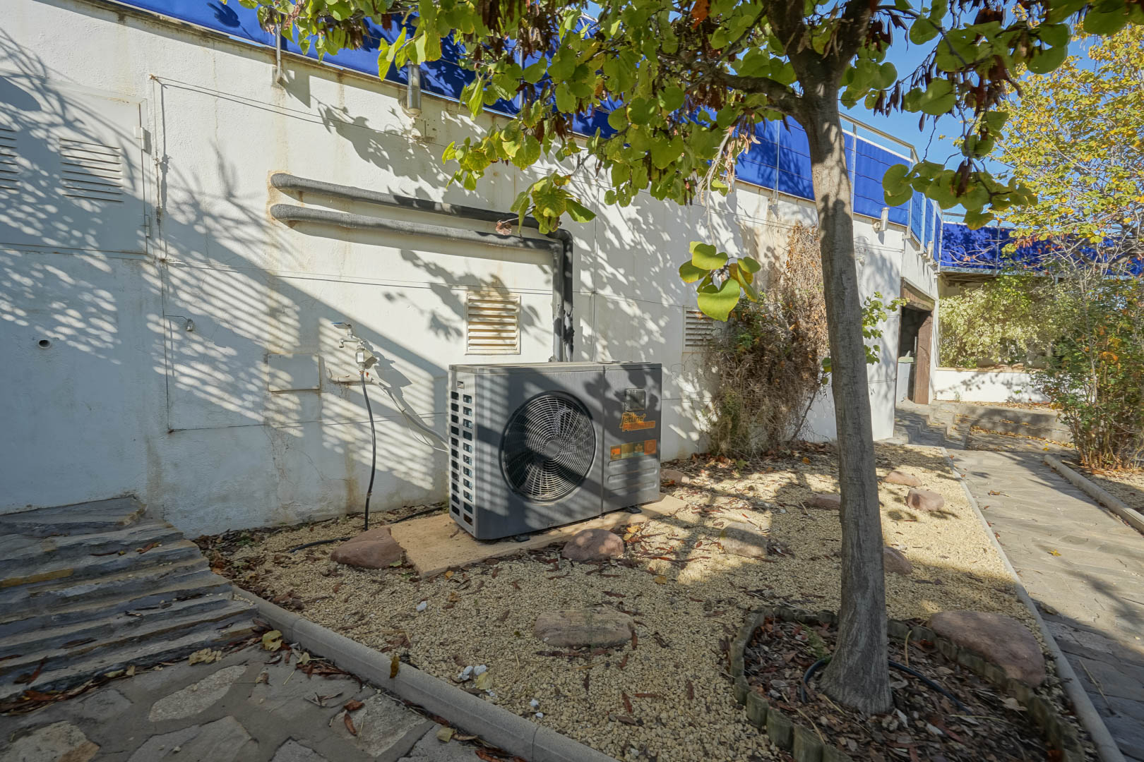 Exterior technical area featuring a Jetline Premium heat pump for the pool, situated on a gravel bed adjacent to stone pathways and garden trees.