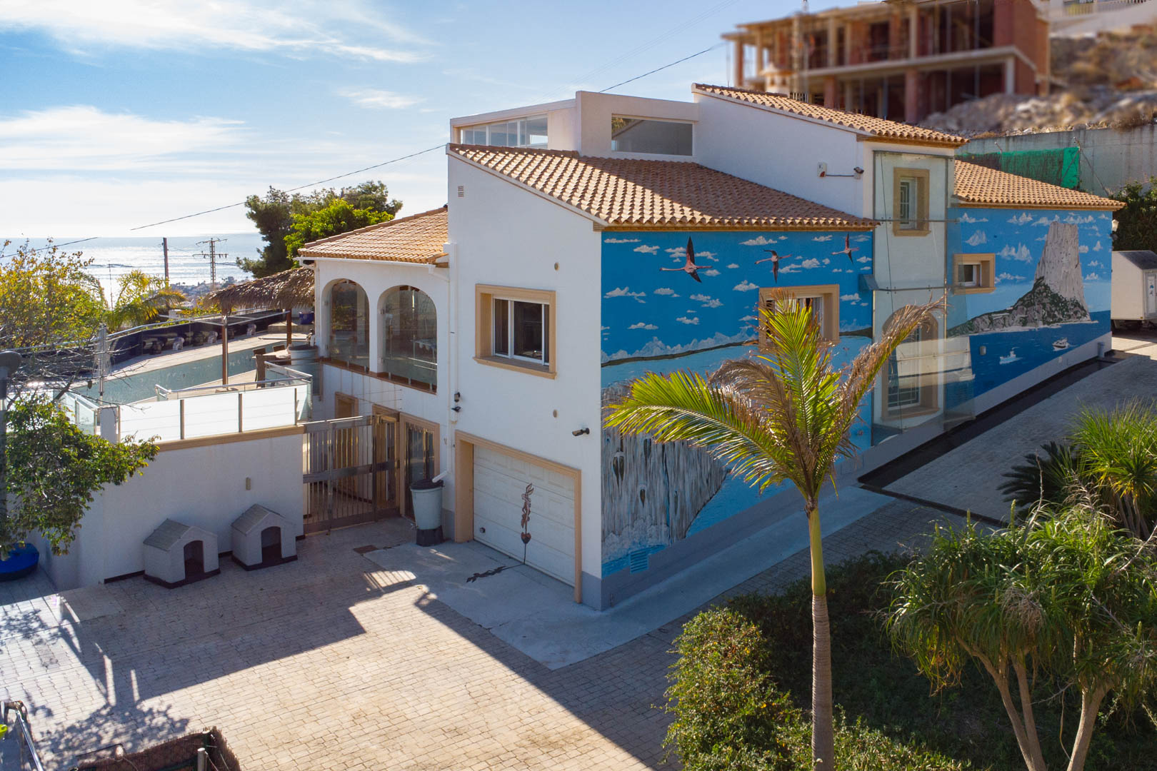 Exterior view featuring a distinctive mural of the Peñón de Ifach, a private garage, and glass-enclosed terraces overlooking the paved entrance.
