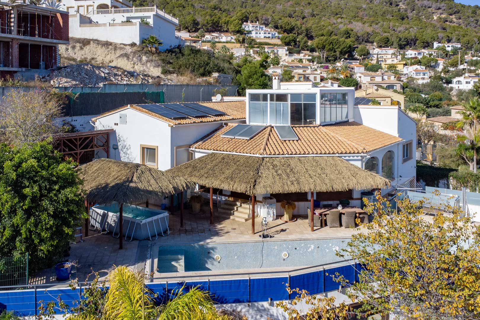 Exterior view of the villa featuring solar panels, thatched-roof pergolas over the terrace, a swimming pool, and a glass-enclosed upper level overlooking the hillside.