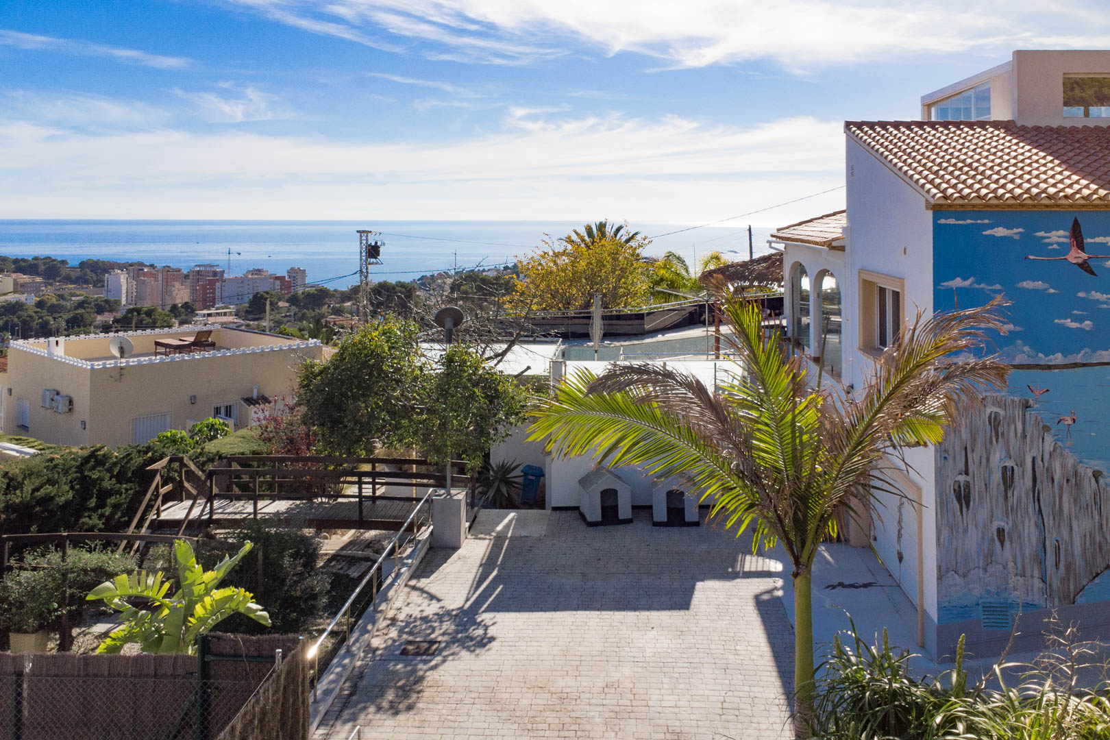Exterior view showing the paved entrance, a flamingo mural on the facade, and panoramic views of the Mediterranean Sea and Calpe skyline.