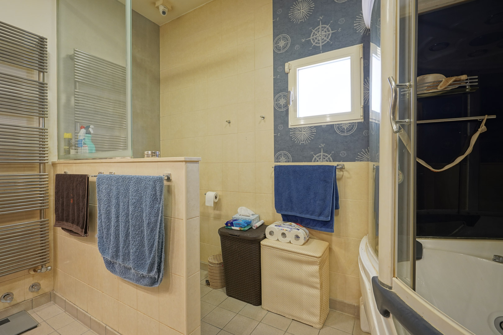 Functional bathroom featuring cream ceramic tiling, a stainless steel towel radiator, and a separate toilet area. A nautical-themed wall accent surrounds the window.