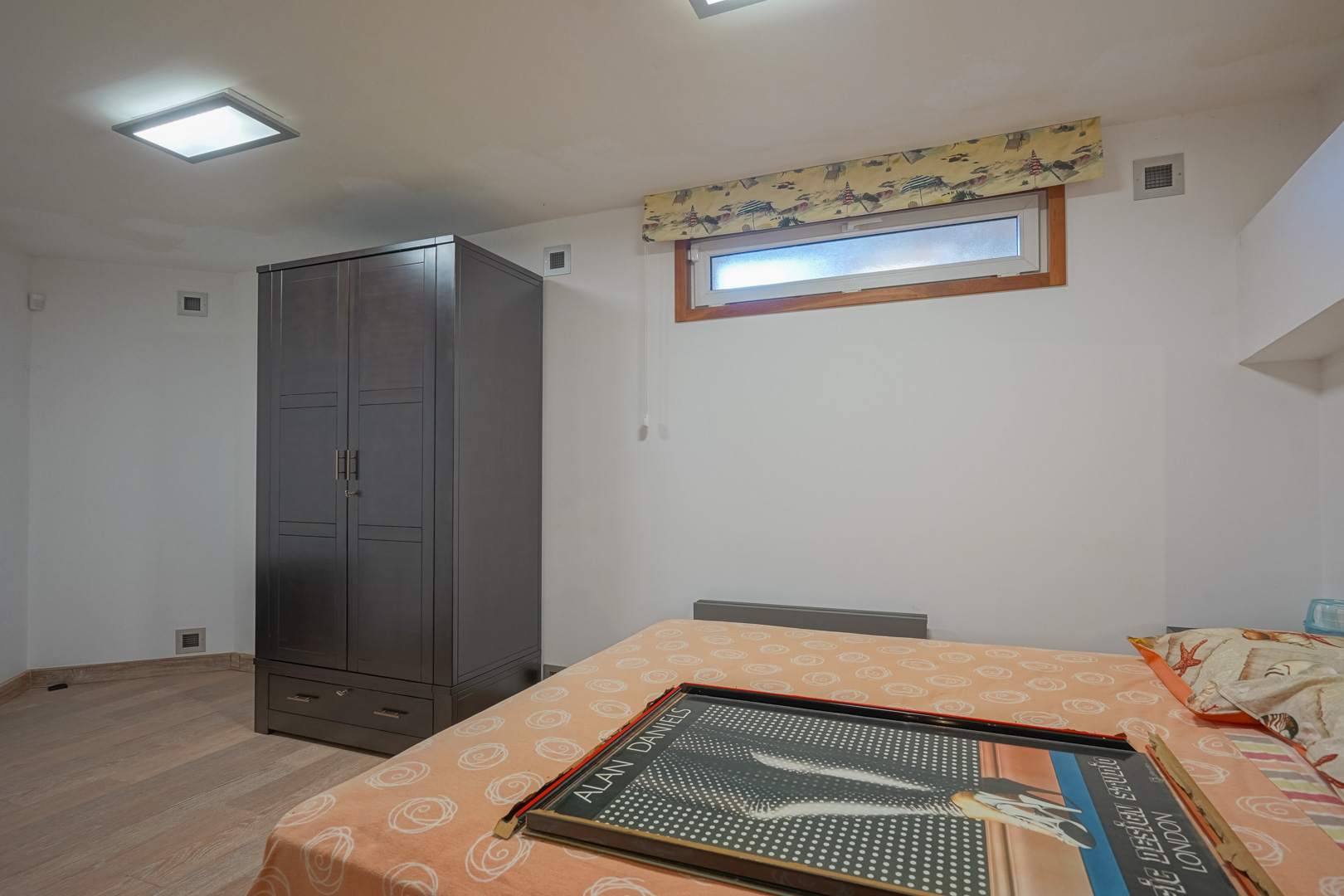 Functional bedroom featuring a dark wood wardrobe, light laminate flooring, and a horizontal window with a patterned blind under recessed ceiling lighting.