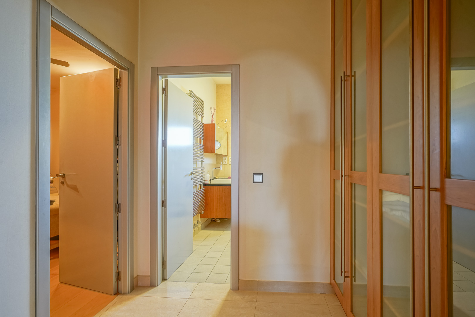 Functional hallway featuring built-in wooden wardrobes with glass panels, connecting a bedroom with hardwood floors to an en-suite bathroom with a towel radiator.