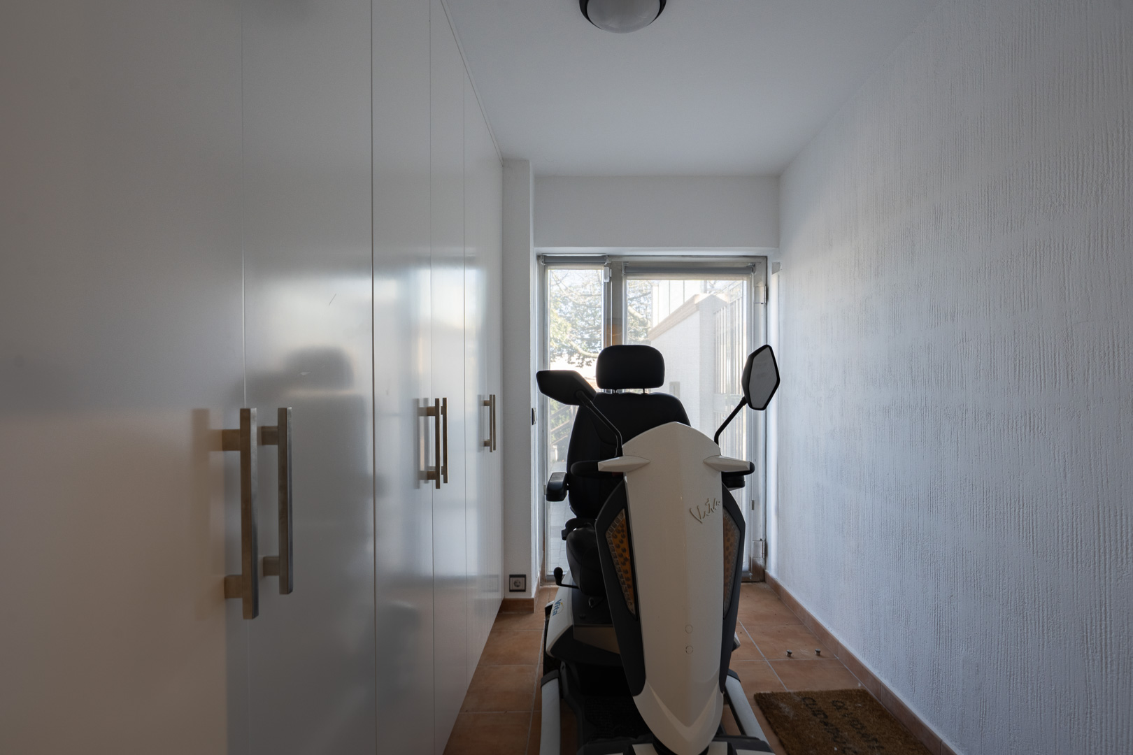Functional hallway featuring large white built-in wardrobes with metallic handles, terracotta tile flooring, and a glass door leading outside.