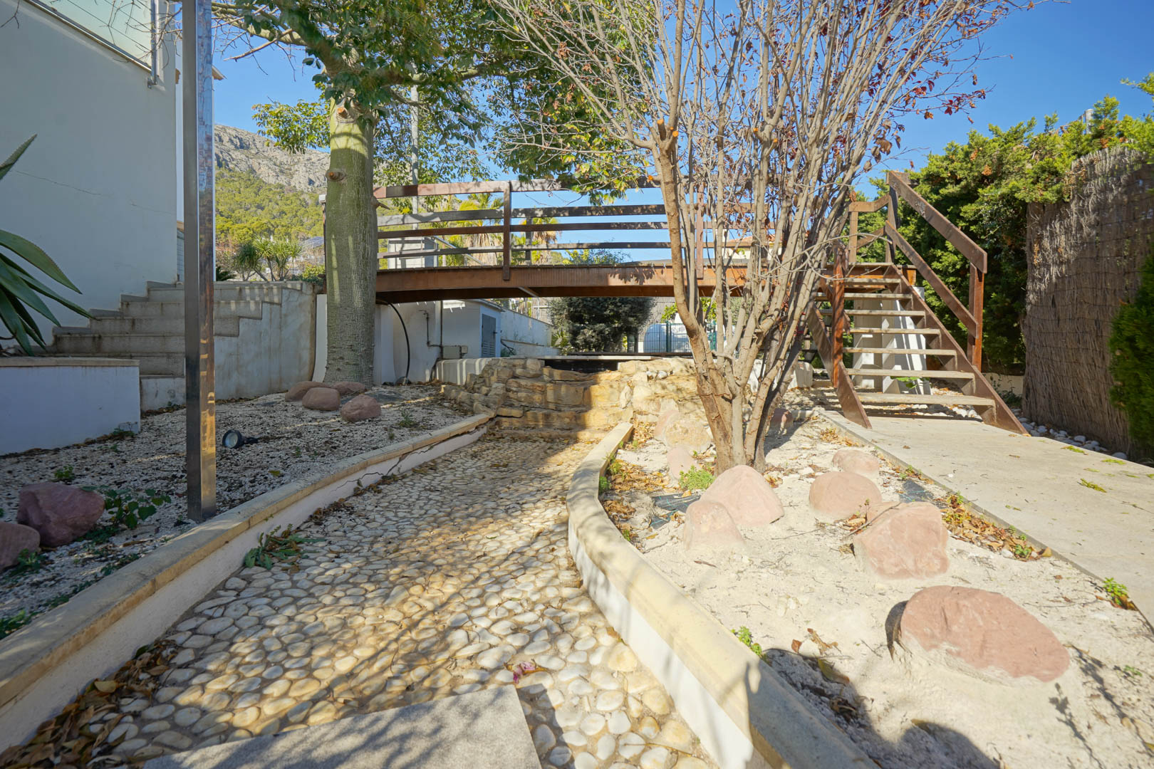 Garden area featuring a wooden bridge over a decorative stone waterfall, a pebble-paved path, and access stairs against the backdrop of the Oltamar mountains.