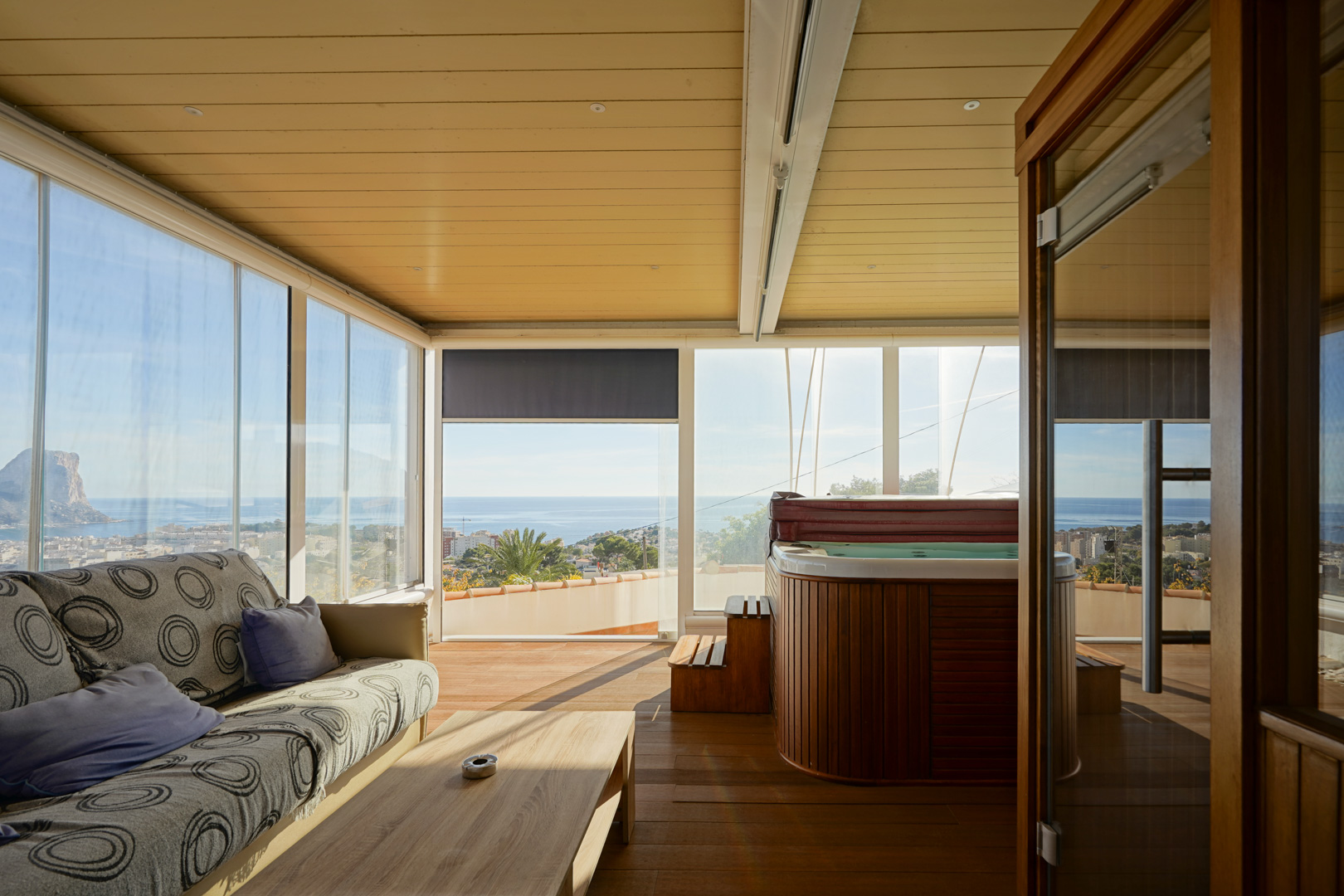 Glazed terrace featuring a wood-clad jacuzzi, lounge area, and panoramic views of the Peñón de Ifach. Includes wooden flooring and a paneled ceiling with recessed lighting.