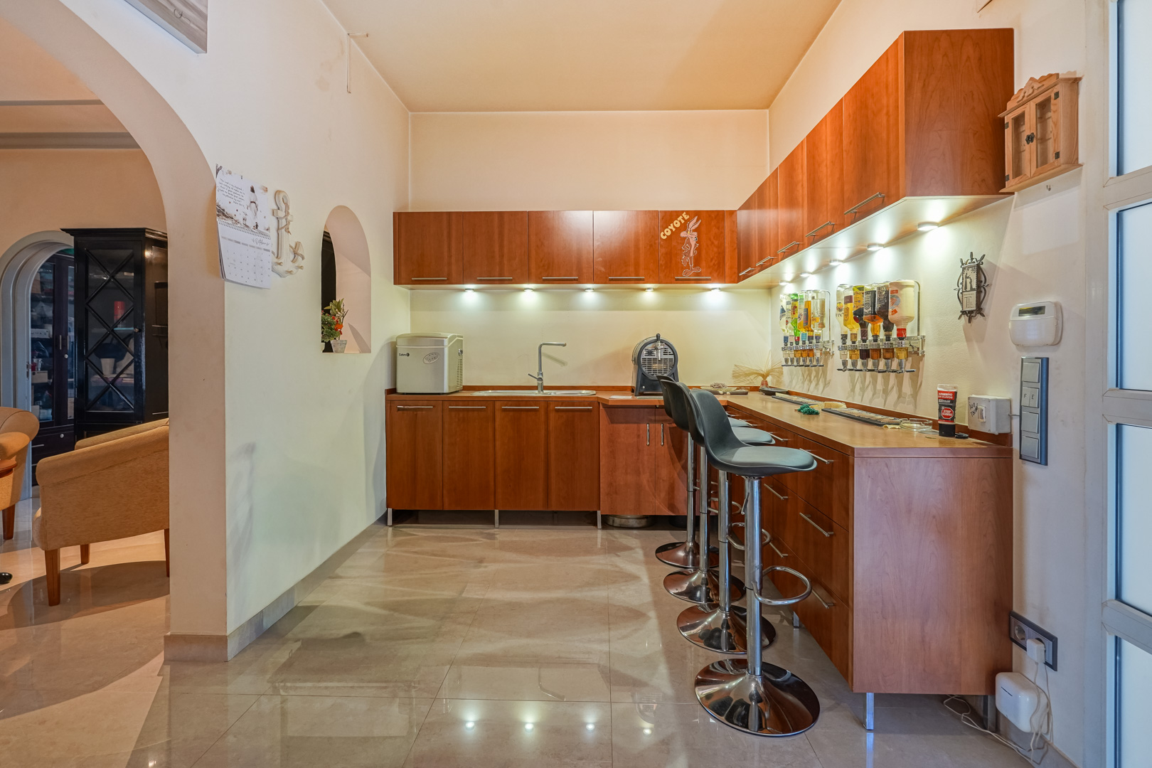 Integrated home bar with wooden cabinetry, a built-in sink, and wall dispensers. Features chrome stools, under-cabinet LED lighting, and polished ceramic flooring.