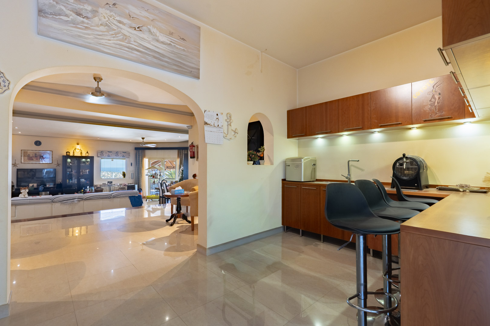 Integrated wooden bar area with high stools and under-cabinet lighting, featuring a wide archway that connects to the main living room and polished tile floors.