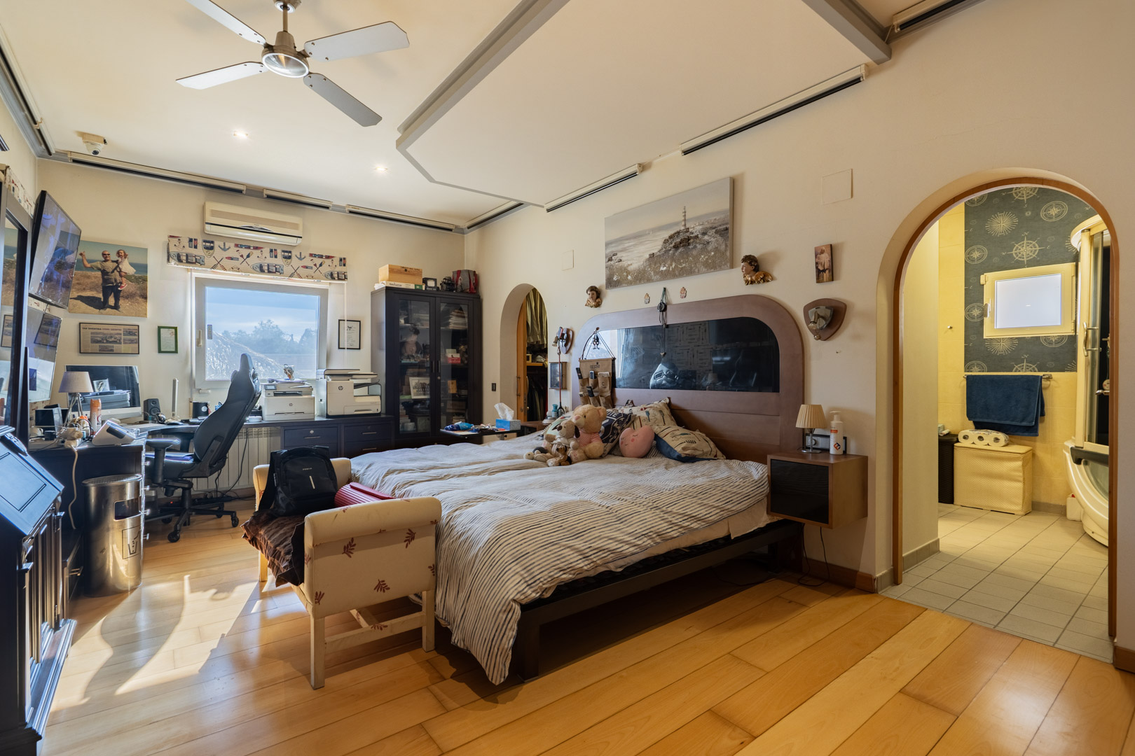 Master bedroom featuring hardwood floors, a dedicated workspace, and an ensuite bathroom. The room includes a ceiling fan and natural light from a side window.