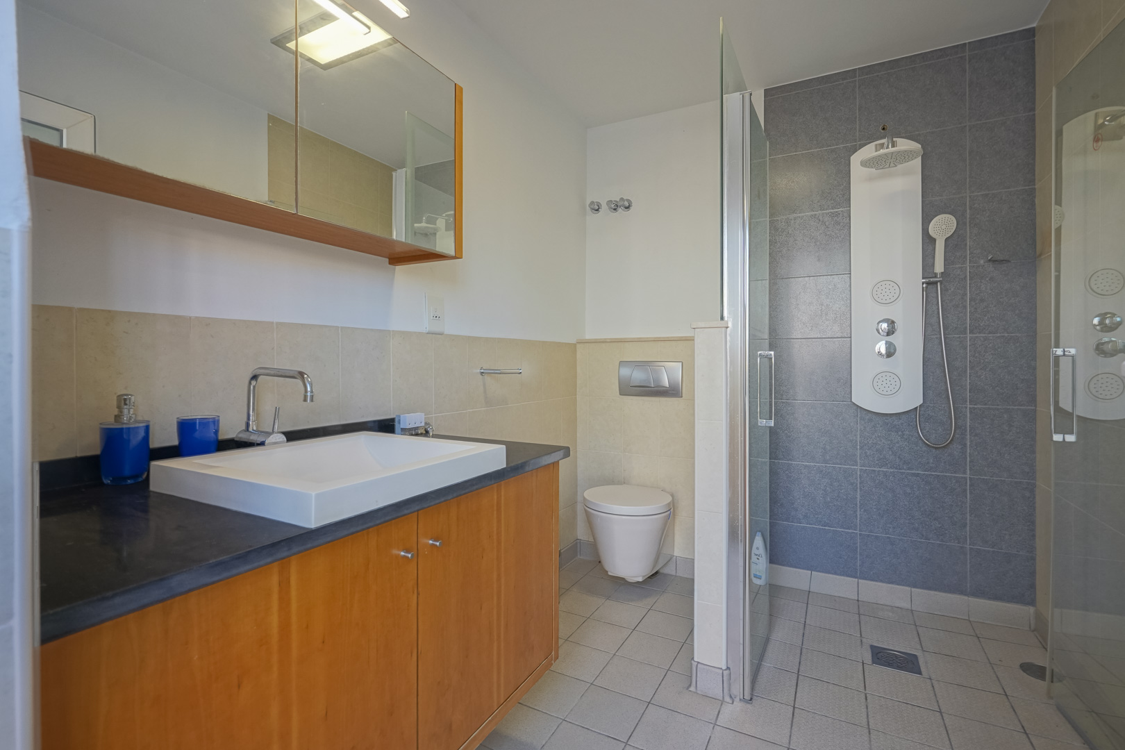 Modern bathroom featuring a wooden vanity with a dark countertop, a walk-in shower with a hydromassage panel, and a mix of neutral beige and grey tiling.