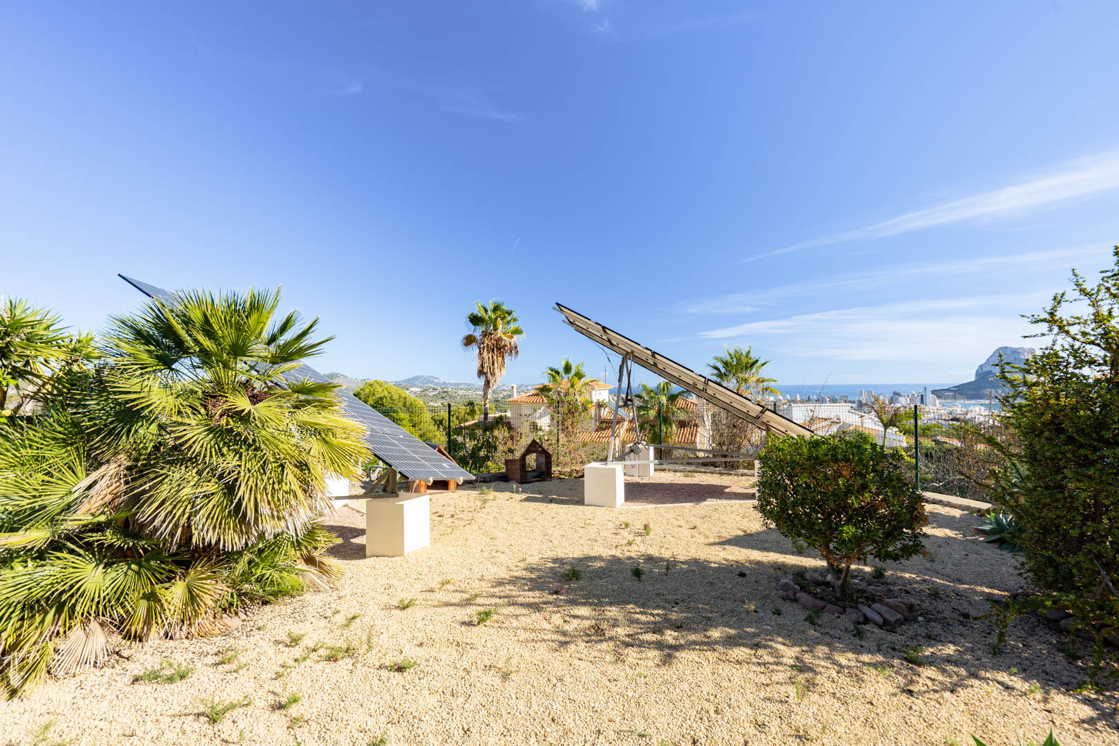 Motorized solar panel trackers installed on concrete bases within a gravel garden, offering clear views of the Peñón de Ifach and Calpe skyline.