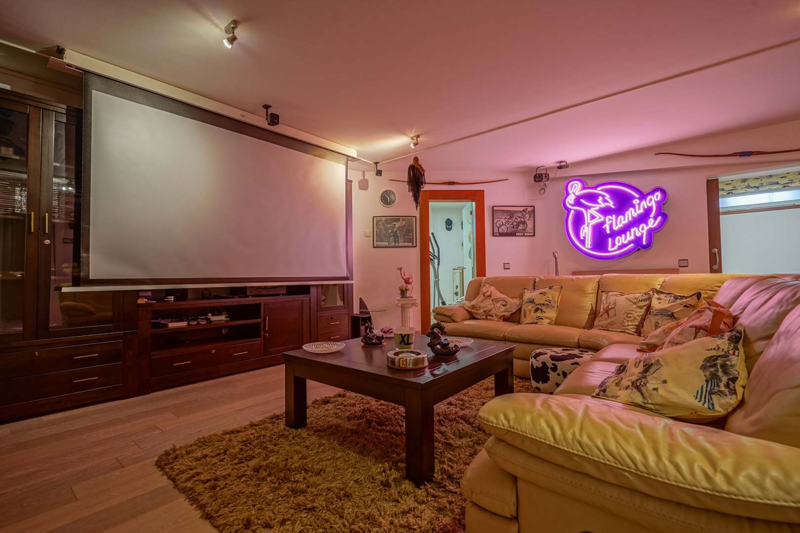 Multimedia room featuring a large projection screen, dark wood cabinetry, and a cream leather corner sofa. Ambient neon lighting and wooden flooring define the space.