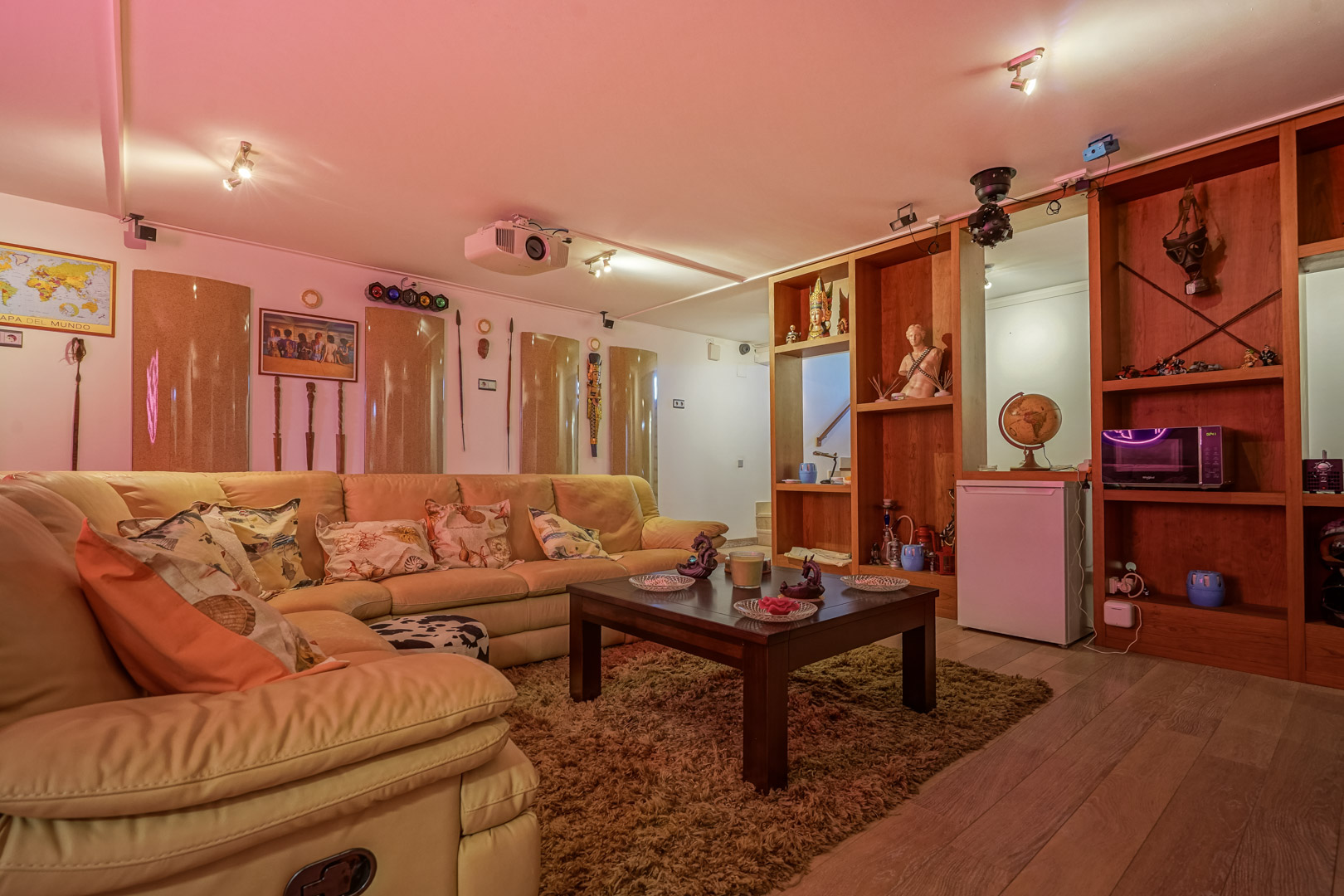 Multimedia room featuring a leather corner sofa, ceiling-mounted projector, disco lighting, and custom wooden shelving with a mini-fridge and microwave.