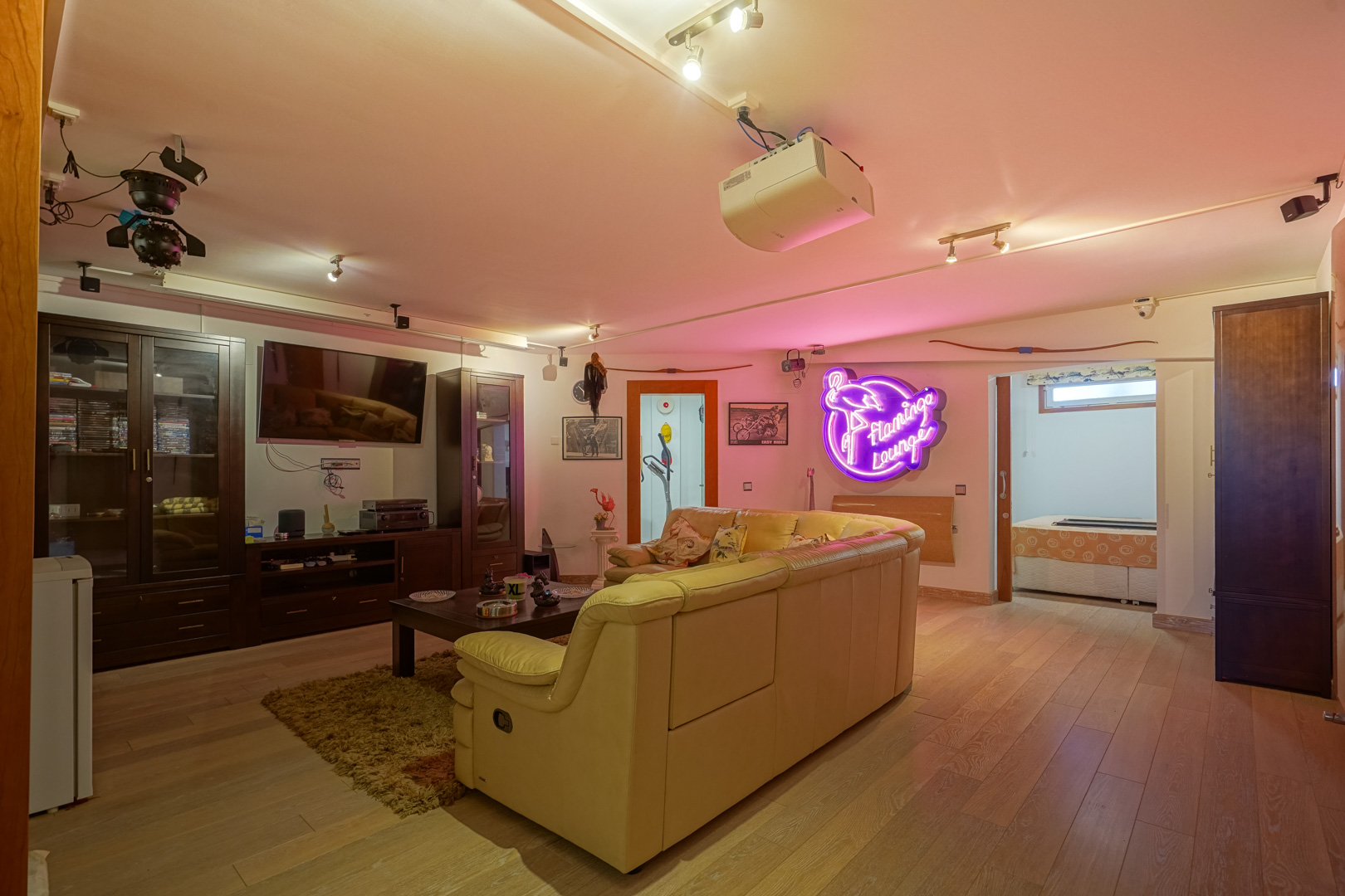 Multimedia room featuring wooden flooring, a ceiling-mounted projector, and a leather sofa. The space includes dark wood cabinetry, track lighting, and a neon lounge sign.
