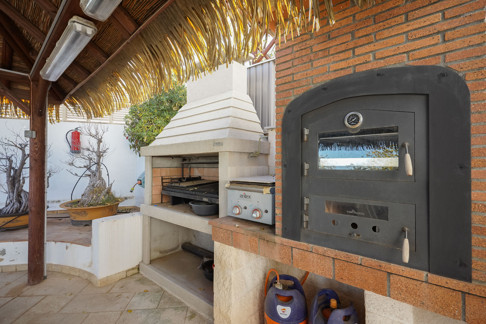 Outdoor cooking station featuring a built-in Tromen oven, Argentine grill, and gas plancha under a rustic wooden pergola with a thatched roof.