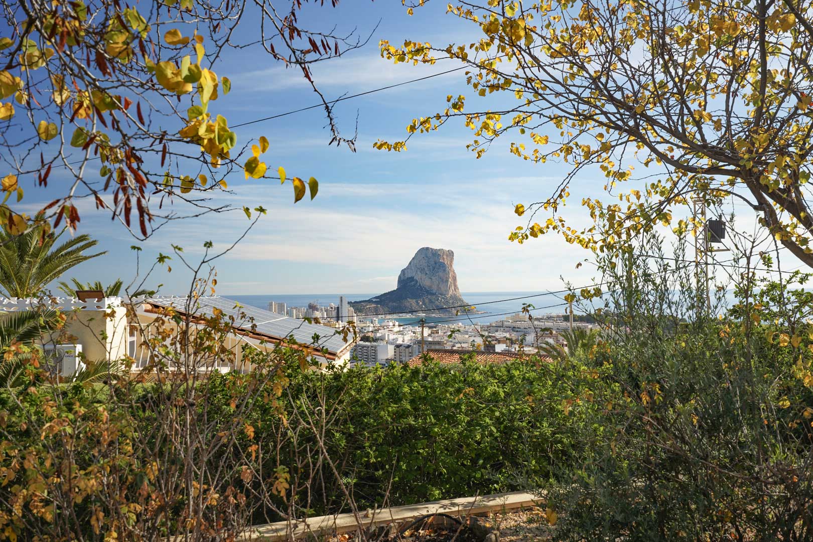 Panoramic view of the Peñón de Ifach and Calpe skyline from the estate, featuring the roof-mounted solar panel system and lush surrounding Mediterranean foliage.