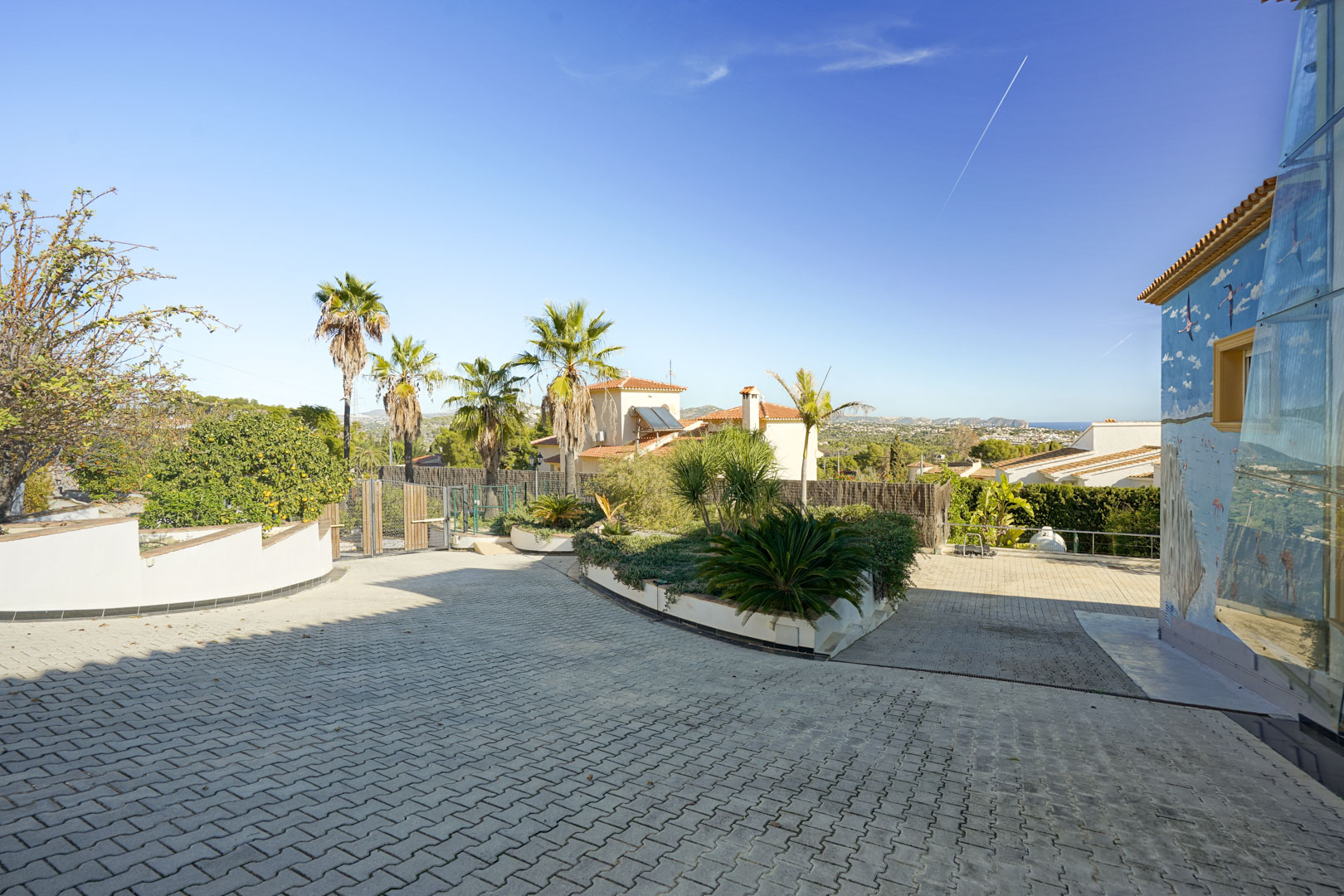 Paved entrance driveway with interlocking stones, featuring a landscaped garden island, a decorative wall mural, and distant views of the Mediterranean coastline.