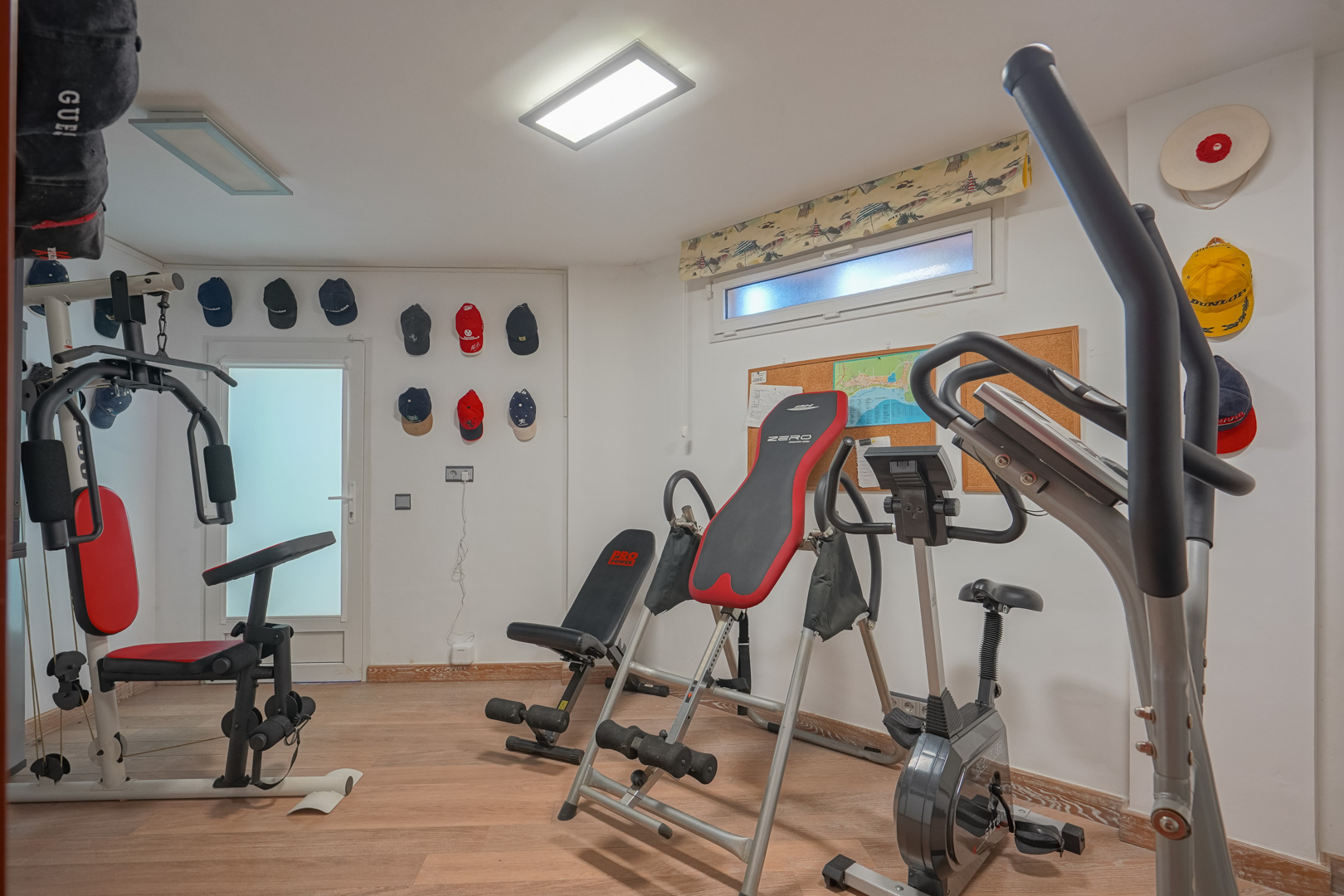 Private gym featuring wooden flooring, equipped with a multi-station machine, stationary bike, and inversion table under LED panel lighting.