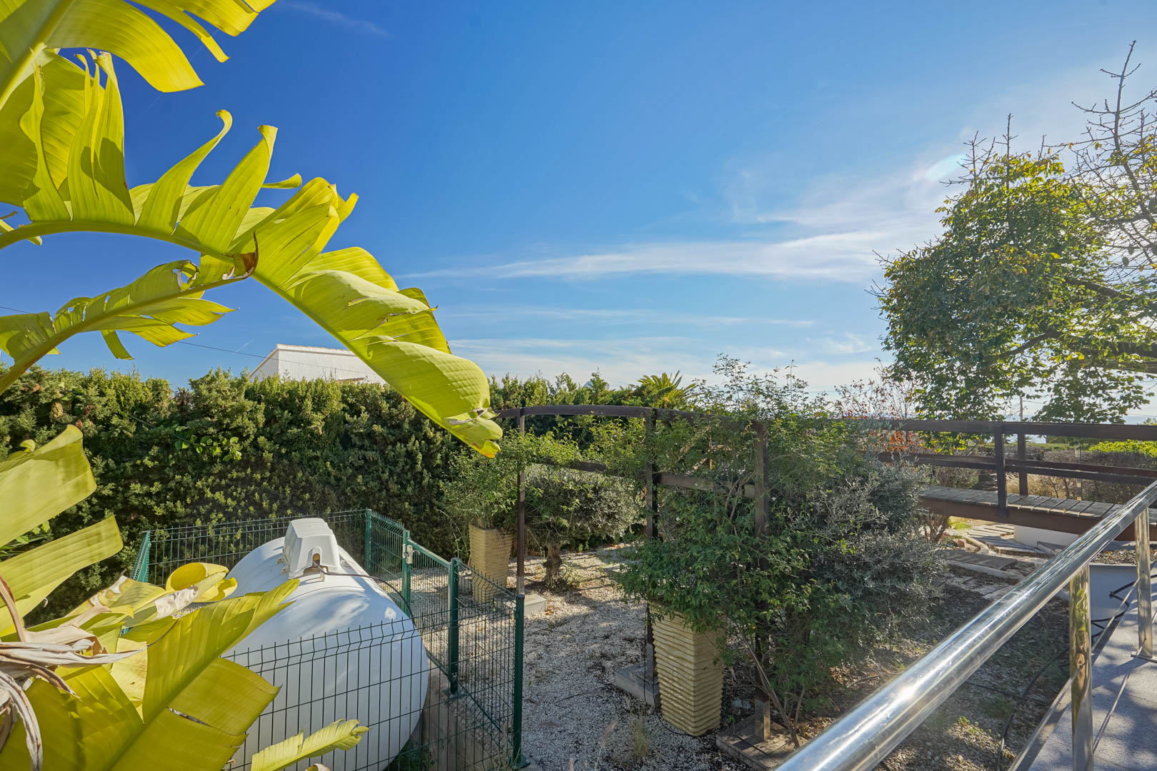 Side garden area featuring a fenced utility tank, a wooden walkway, and tropical foliage under a clear blue sky.
