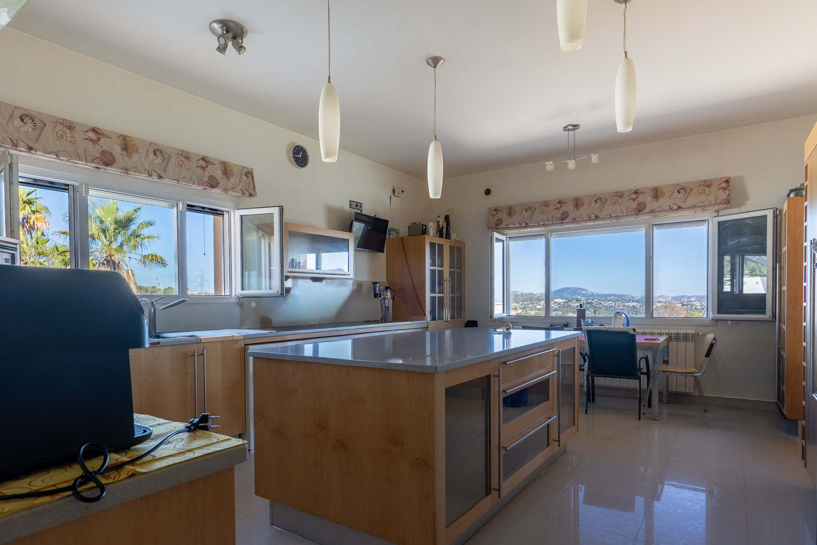 Spacious kitchen featuring a large central island with wooden cabinetry and grey countertops. Large windows provide panoramic views of the coast and mountains.