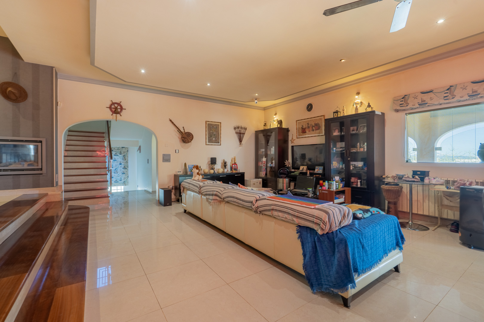 Spacious living area featuring polished tile flooring, a recessed tray ceiling with spotlights, and an arched entrance leading to a wooden staircase and integrated fireplace.