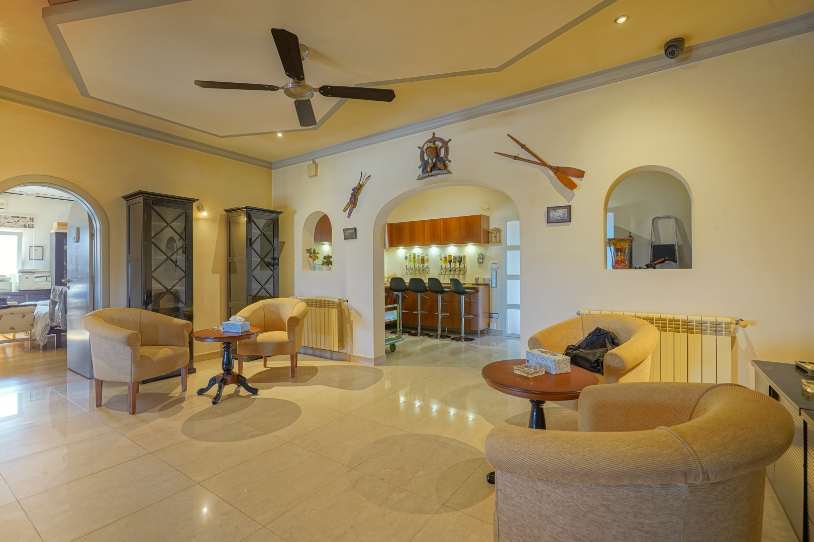 Spacious lounge with a ceiling fan and tiled floors, featuring an integrated bar area visible through arched openings with nautical-themed decor.