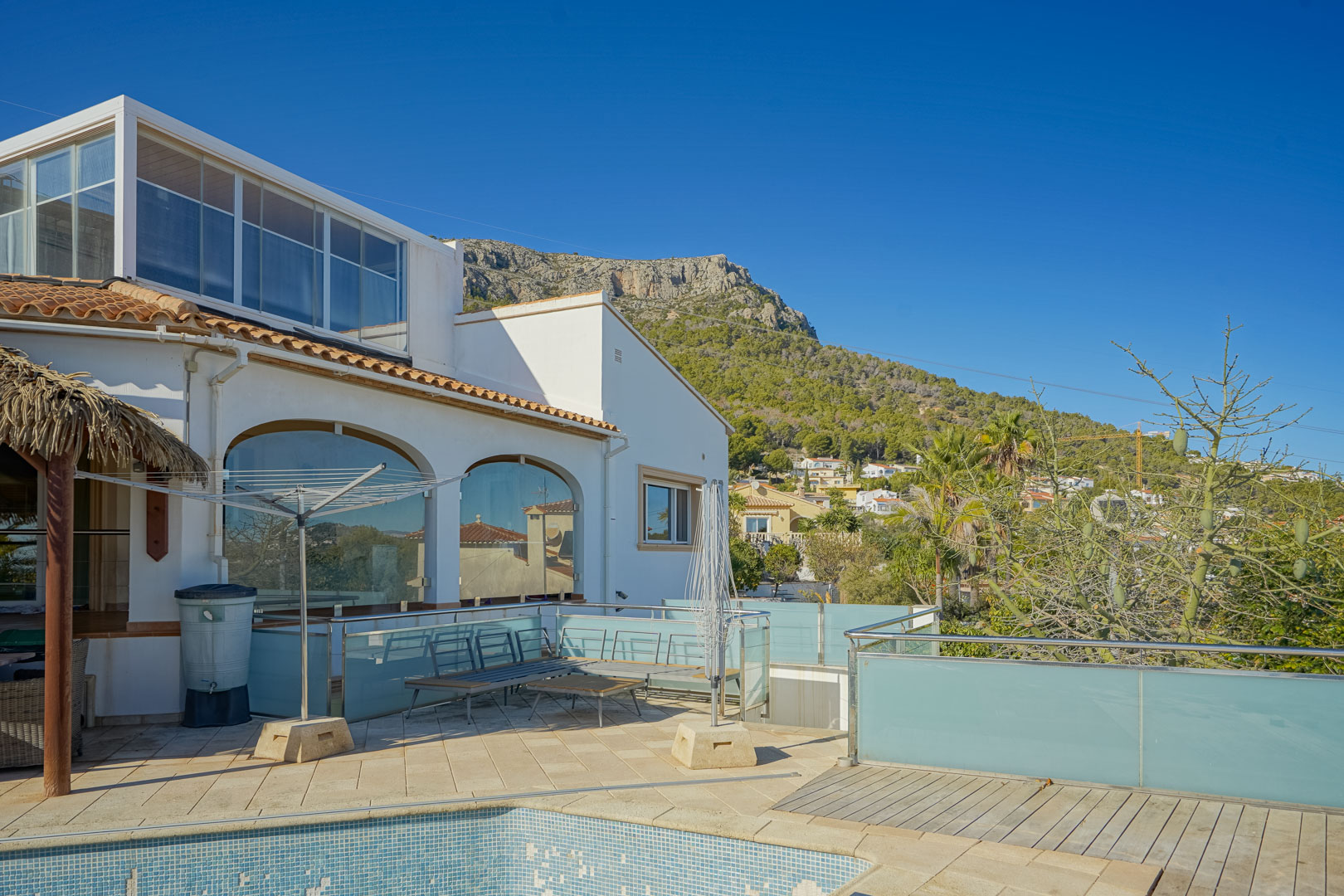 Villa exterior featuring a tiled terrace with glass railings, arched windows, and a glass-enclosed upper level, all set against the backdrop of the Oltamar mountains.