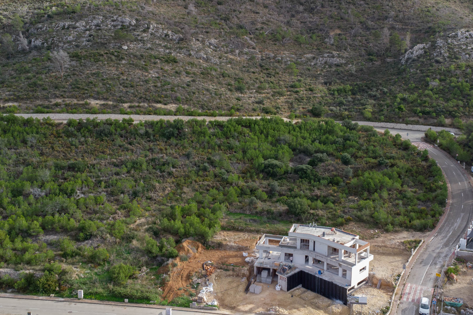 Aerial view of a concrete villa structure under construction in Monte Olímpico, Jávea, bordered by Mediterranean hillside and paved access roads.