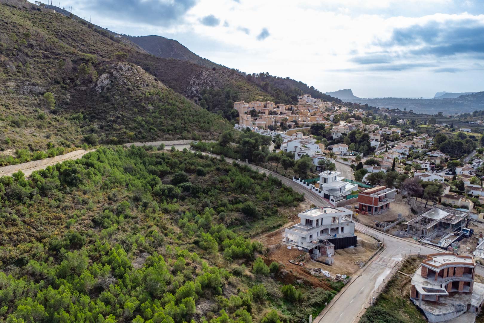 Elevated view of the plot in Monte Olímpico, featuring paved access, neighboring villas under construction, and a lush green hillside overlooking the valley.