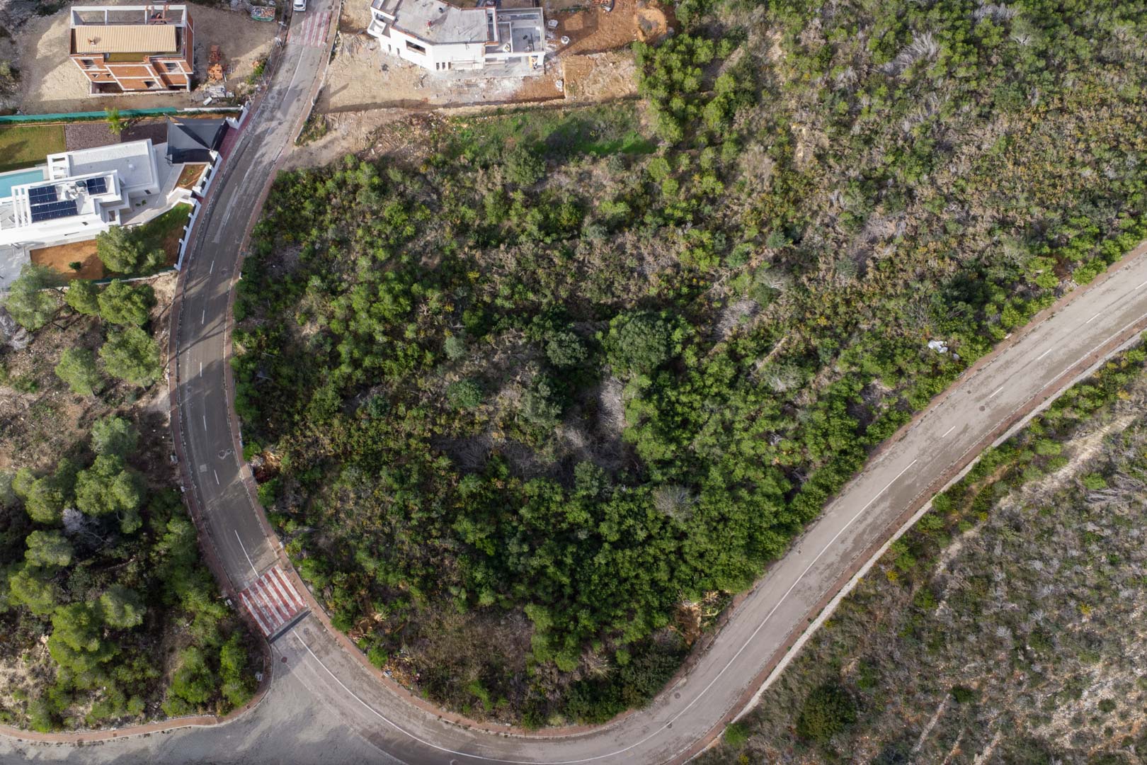Overhead aerial view of the corner urban plot, featuring paved road access with sidewalks and natural Mediterranean shrubland in Monte Olímpico.