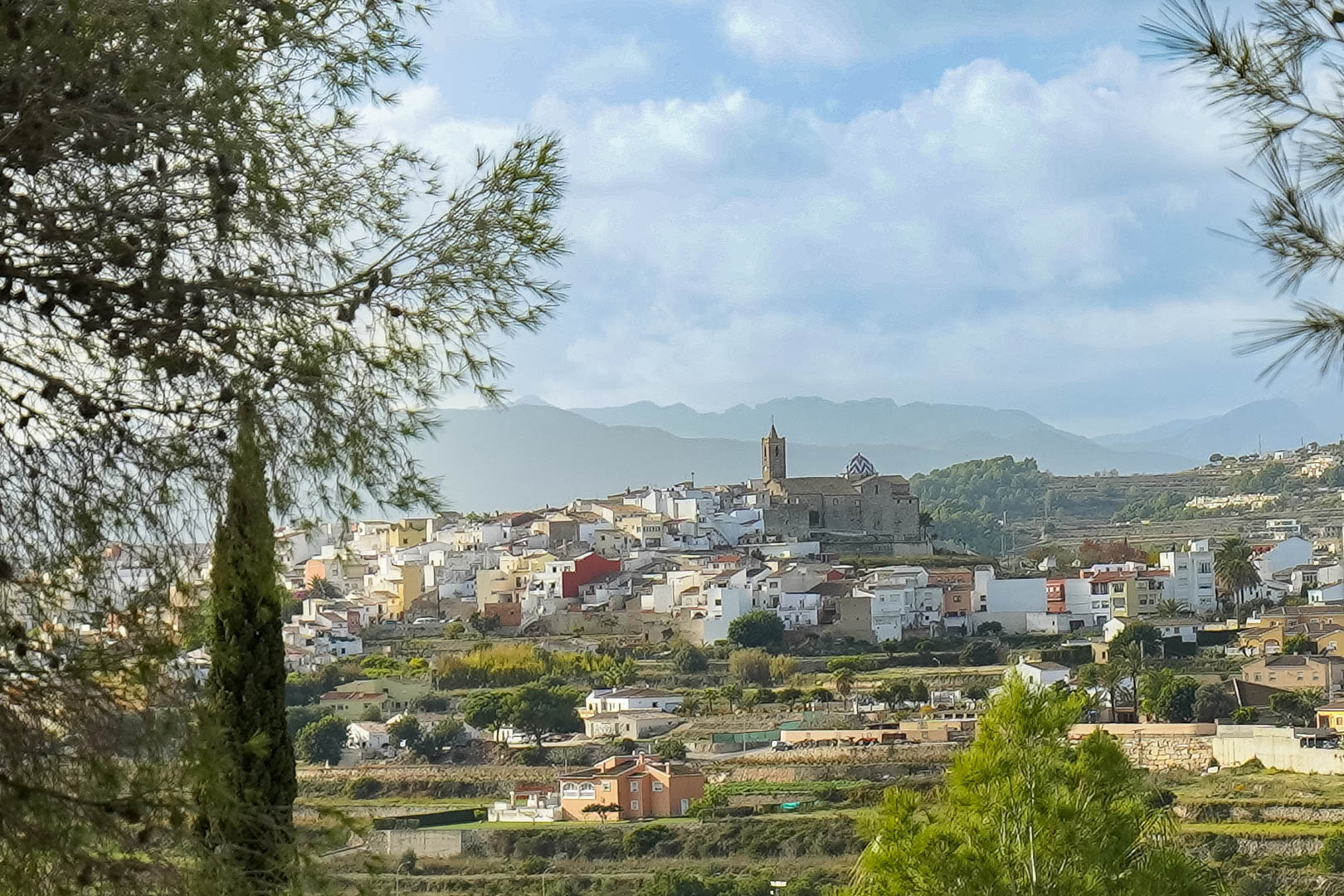 Panoramic views of Benitachell's historic center and church, framed by Mediterranean pines with the Sierra de Bernia mountains in the distance.
