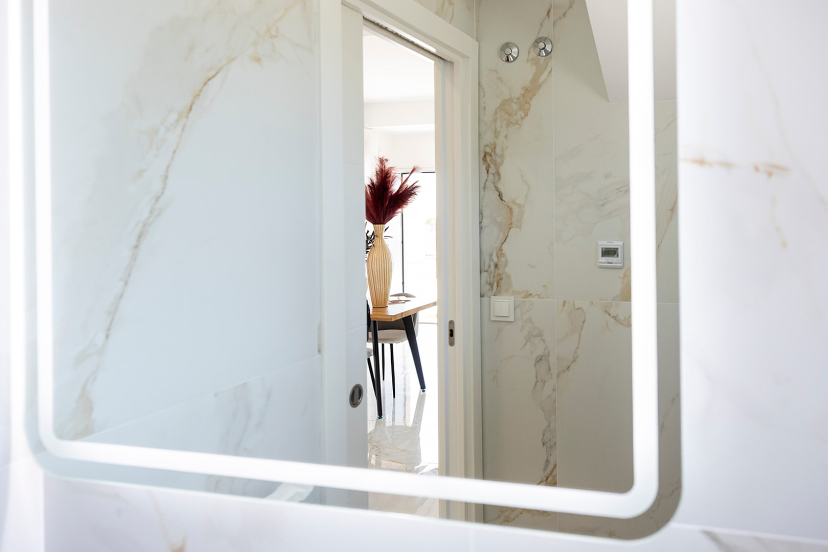 Bathroom featuring marble-effect wall tiles and an LED-backlit mirror, offering a view towards the dining area through a white doorway.