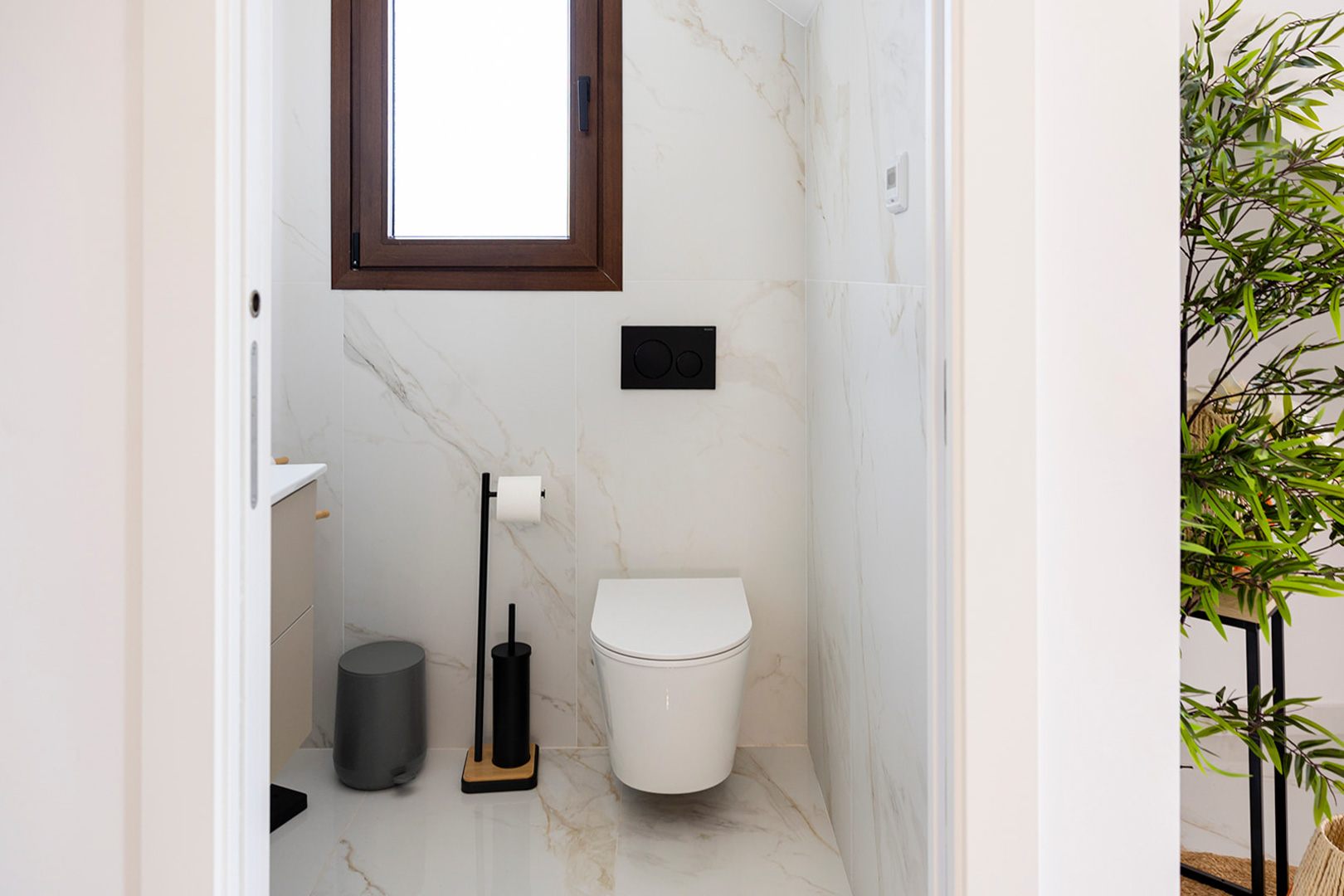 Modern guest bathroom featuring marble-effect tiling, a wall-hung toilet with black hardware, and natural light from a wood-framed window.