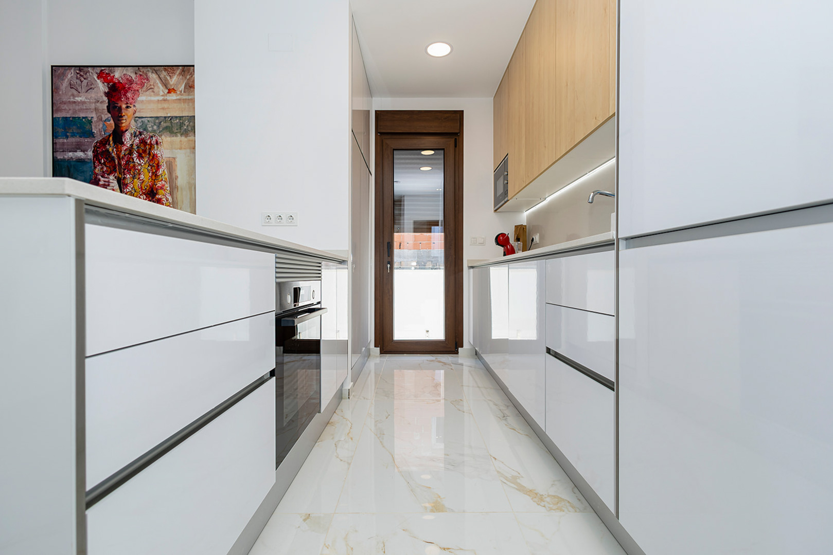 Modern kitchen featuring high-gloss white cabinetry, wooden upper units, and marble-effect porcelain flooring. It includes integrated appliances and a glass door to the exterior.