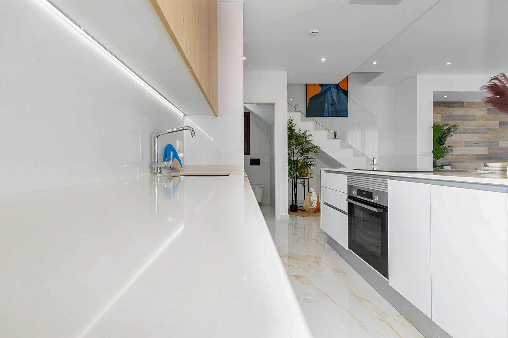 Modern kitchen featuring white countertops, under-cabinet LED lighting, and a central island with an integrated oven. A glass-railed staircase is visible beyond.