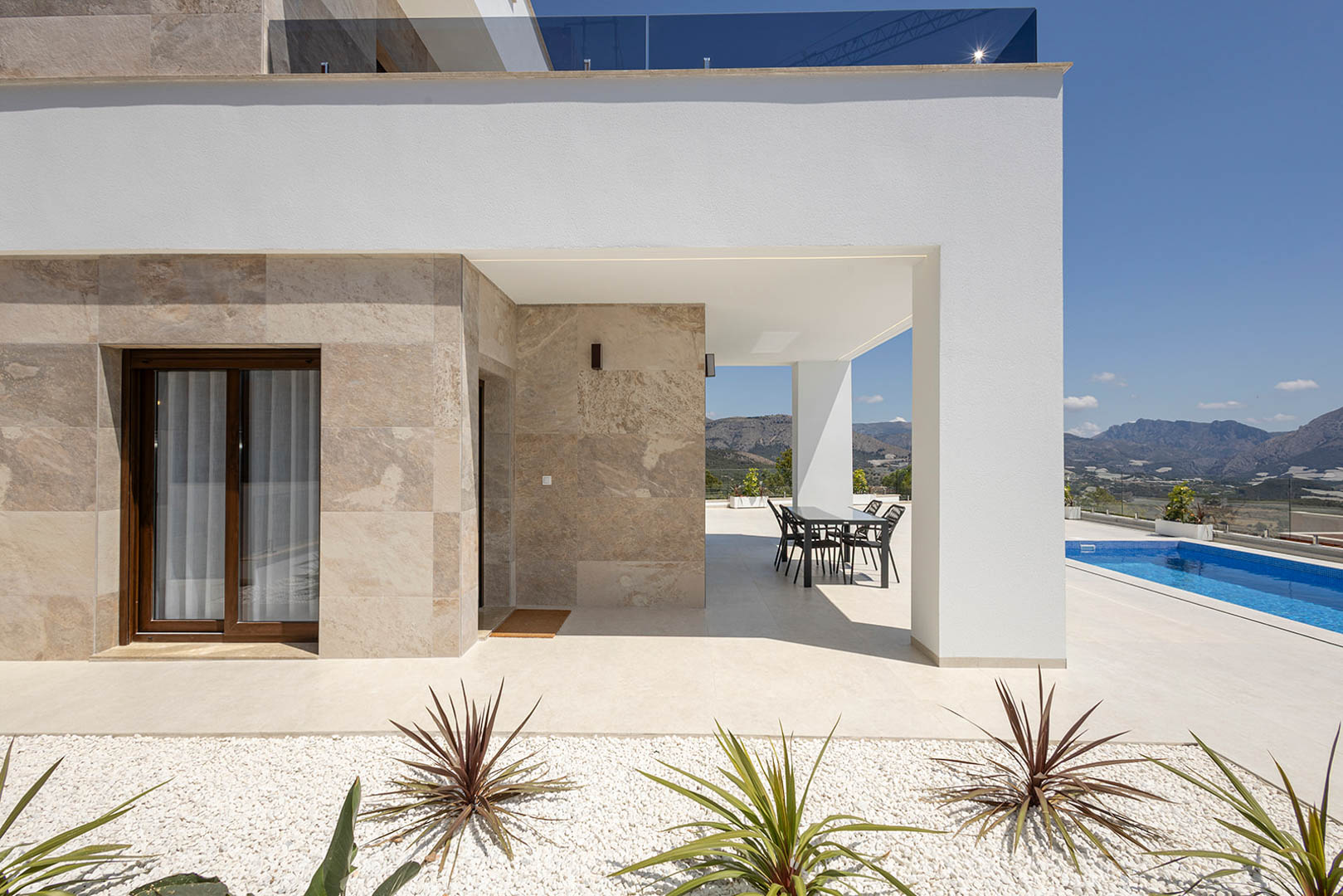 Modern villa exterior featuring natural stone cladding, a covered dining terrace, private swimming pool, and panoramic mountain views.