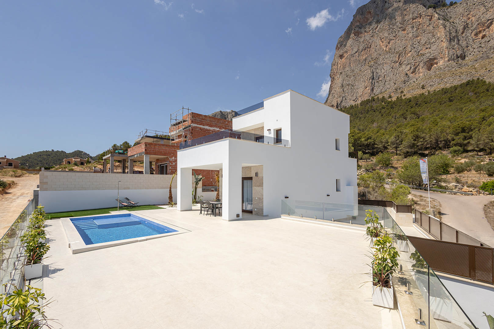 Modern white villa exterior featuring a rectangular blue-tiled pool, expansive porcelain terrace, and glass railings set against a dramatic mountain backdrop.