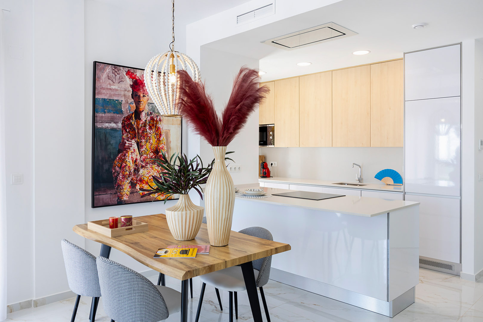 Open-plan kitchen with a white island and light wood cabinets, integrated with a dining area featuring a wooden table, patterned chairs, and a beaded pendant light.