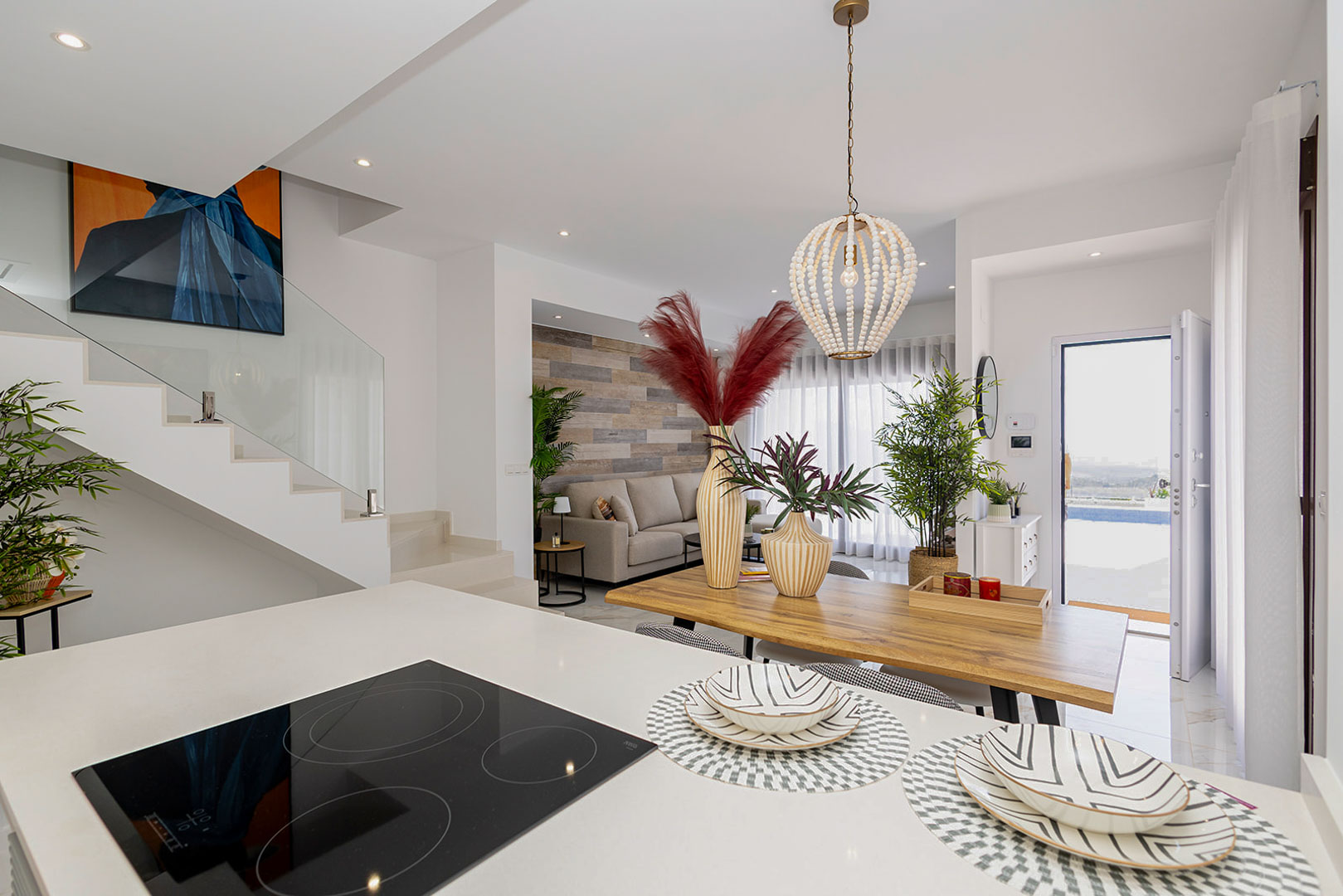 Open-plan living area featuring a kitchen island with induction hob, wooden dining table, and a modern staircase with glass balustrade.