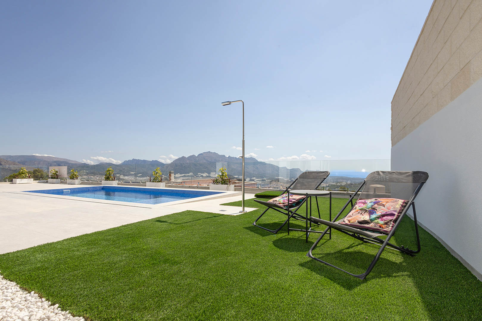 Outdoor terrace featuring a blue mosaic pool, artificial lawn with sun loungers, and a stainless steel shower. Glass railings offer unobstructed mountain and sea views.