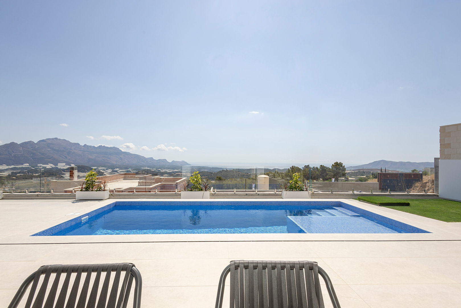 Private blue-tiled swimming pool with integrated steps, featuring a light stone terrace and glass railings overlooking the sea and mountain landscape.
