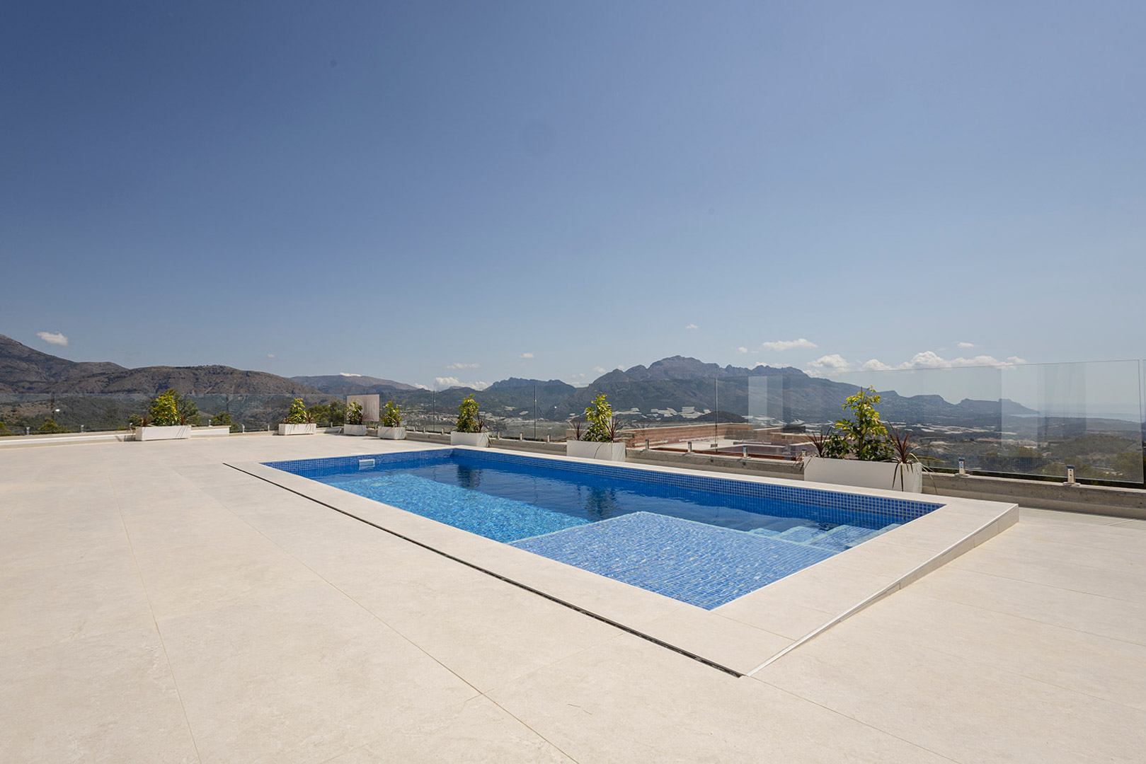 Private pool with blue mosaic tiling set in a spacious light-toned terrace, featuring frameless glass railings and panoramic mountain and sea views.