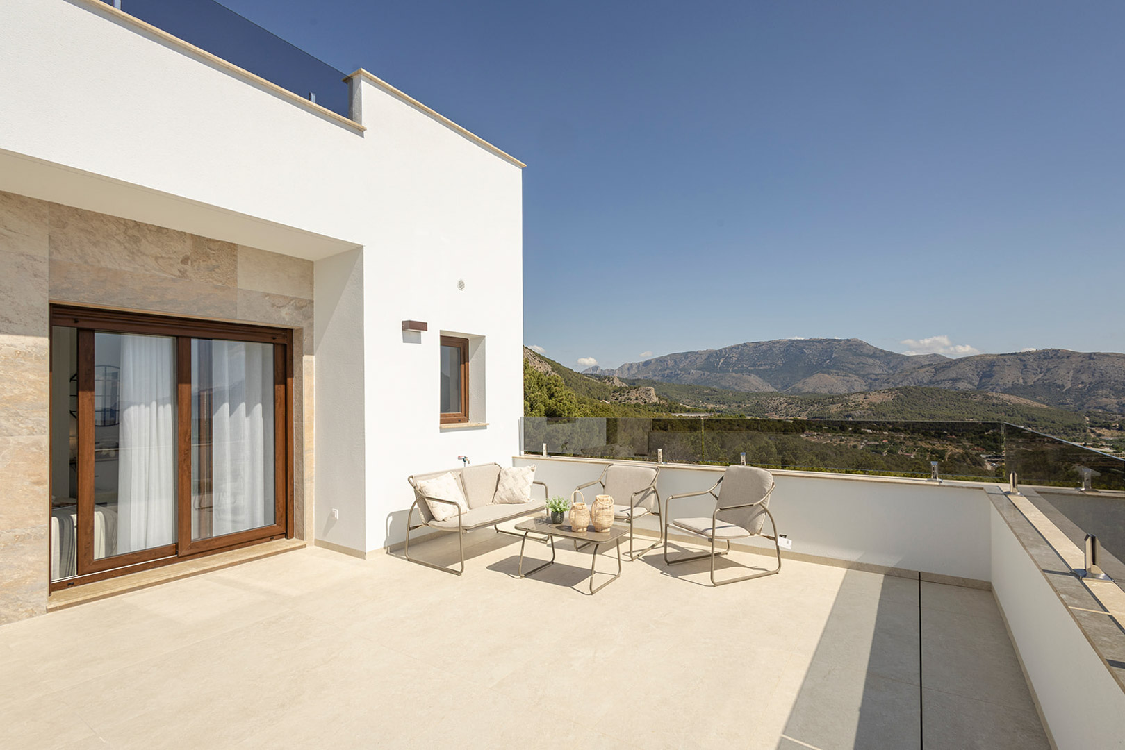 Upper terrace featuring glass railings, outdoor lounge furniture, and panoramic mountain views. White facade with stone accents and light ceramic flooring.