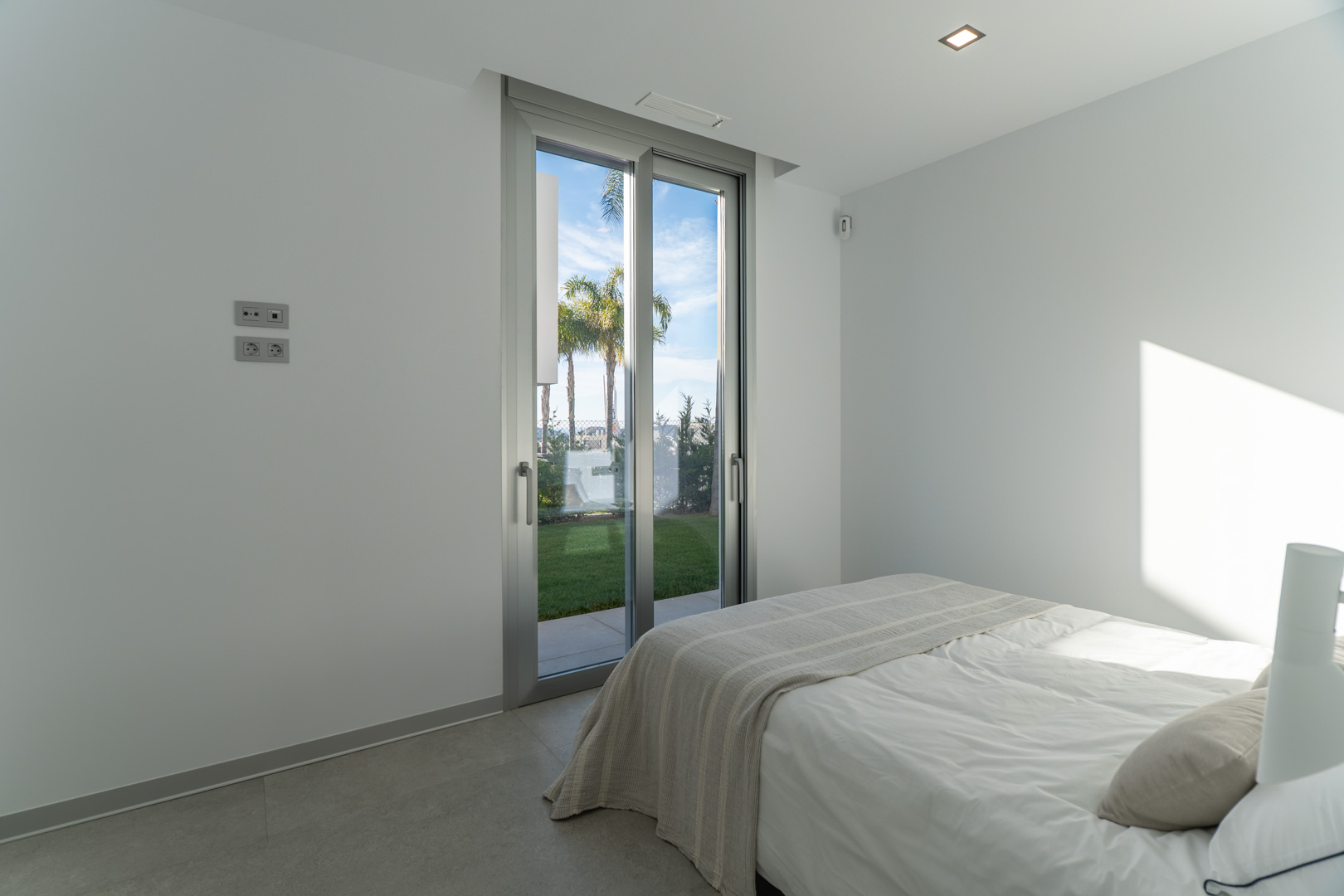 Bright bedroom featuring floor-to-ceiling sliding glass doors with garden views, minimalist white walls, and large-format grey floor tiles.